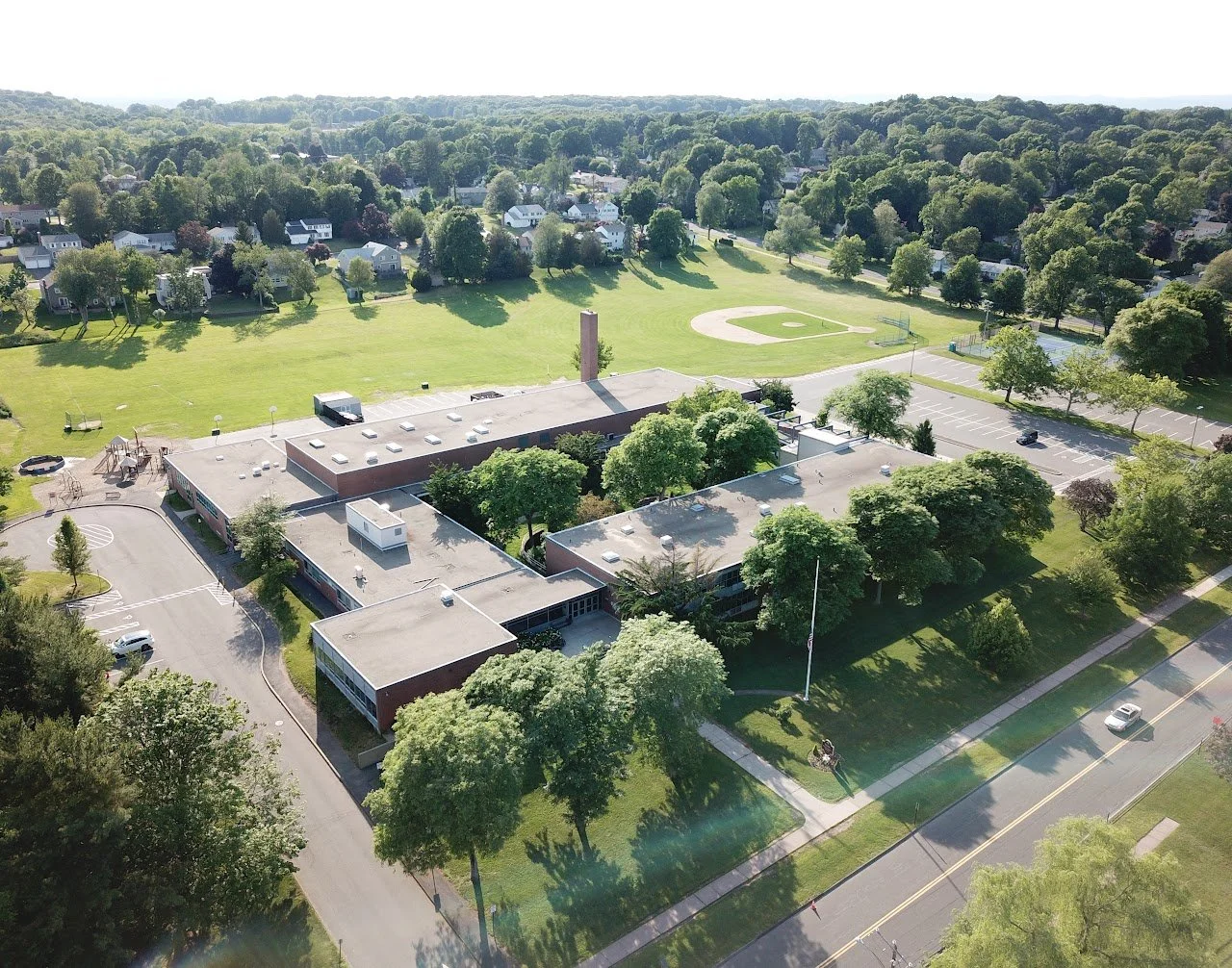 Aerial view of a school campus with multiple flat-roofed buildings, a parking lot, a playground, and a baseball field, surrounded by trees and residential neighborhood.