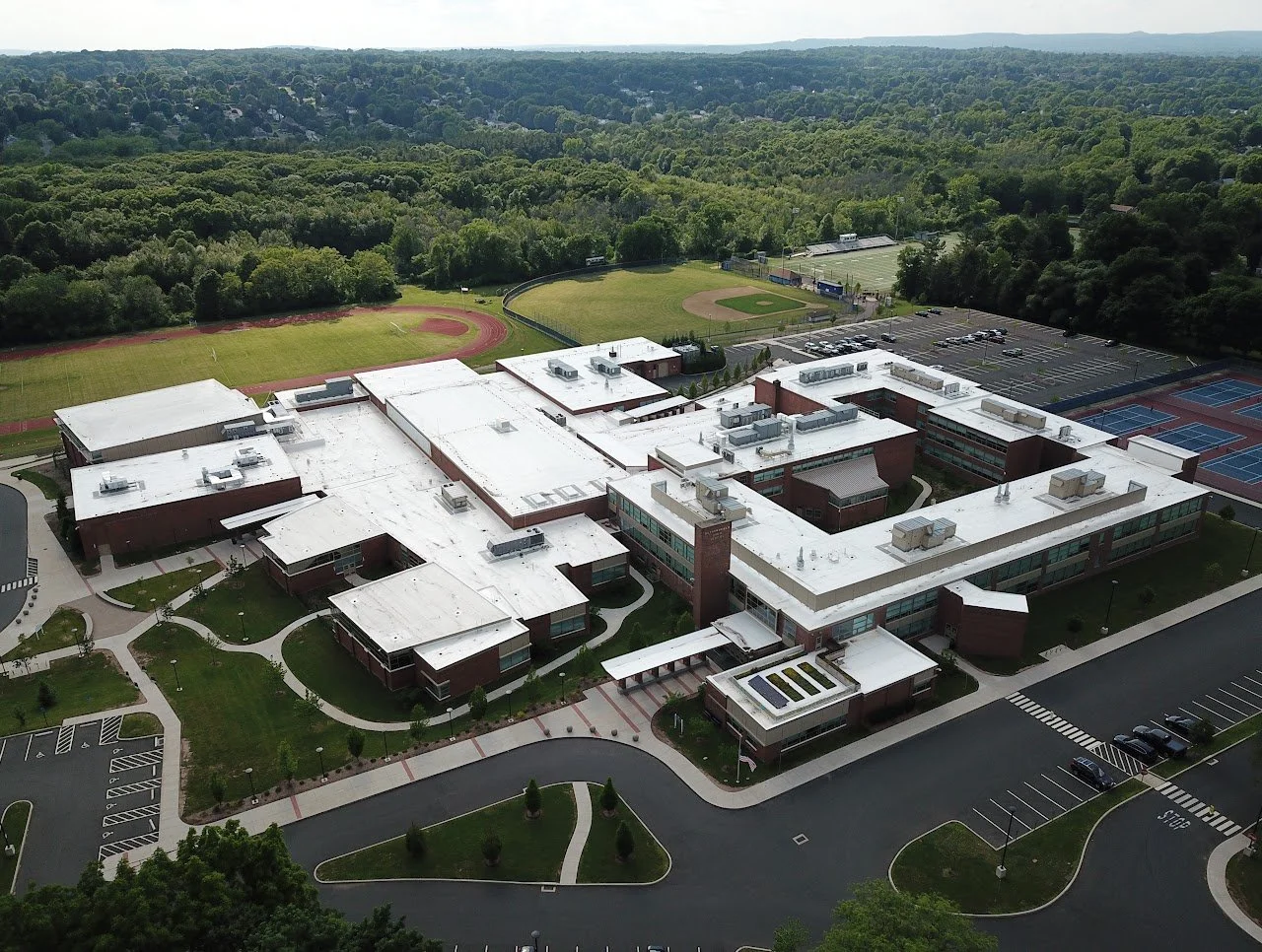 Aerial view of a school campus with a large building, sports fields, parking lot, and surrounding greenery.