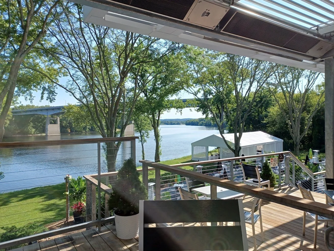 A view from a deck overlooking a river with trees, a bridge in the background, and a white canopy tent set up on the grass.