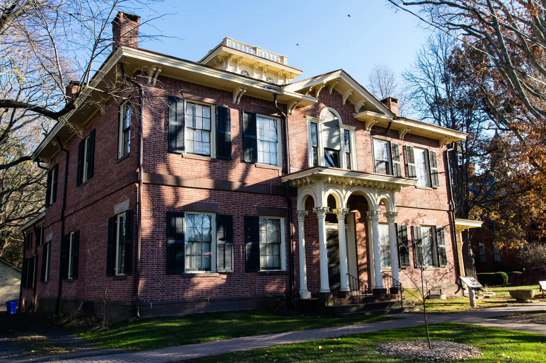 Large historic brick house with black shutters and decorative columns at the front entrance, surrounded by a lawn and leafless trees.