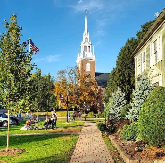A small town park with a paved walkway leading to a church with a tall white steeple, surrounded by trees and American flags under a blue sky.