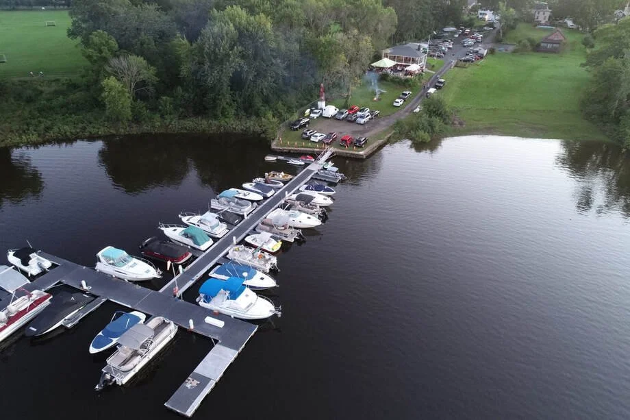 Aerial view of a marina with boats docked at piers on a calm body of water, adjacent to a parking lot and a green park with trees and a building with an outdoor seating area.