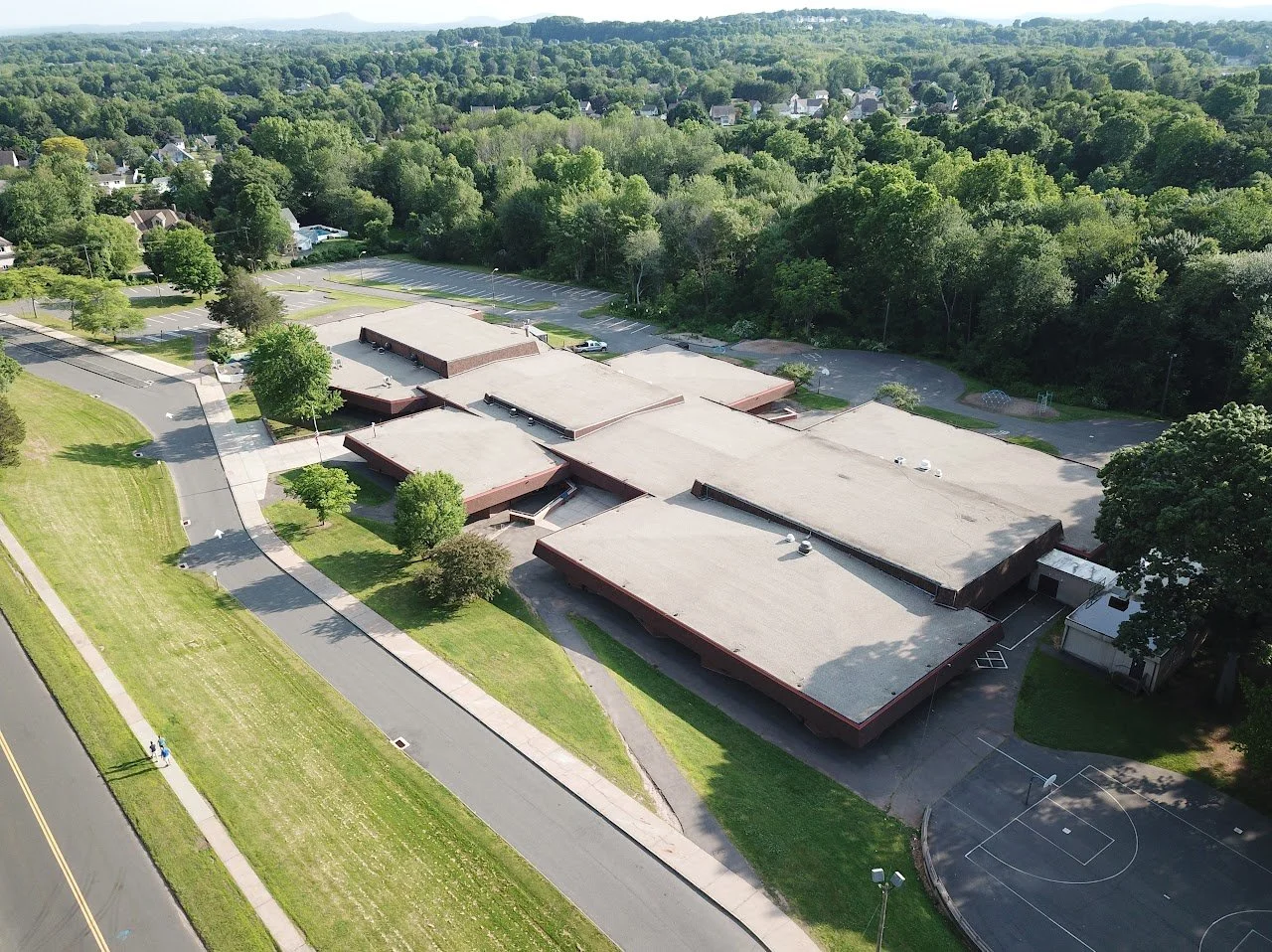 Aerial view of a school building surrounded by parking lots, green trees, and a sports court, with a residential area and forest in the background.