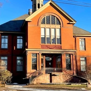 Brick building with a large arched window and smaller windows, featuring a decorative entrance with stairs and a railing, under a clear blue sky.