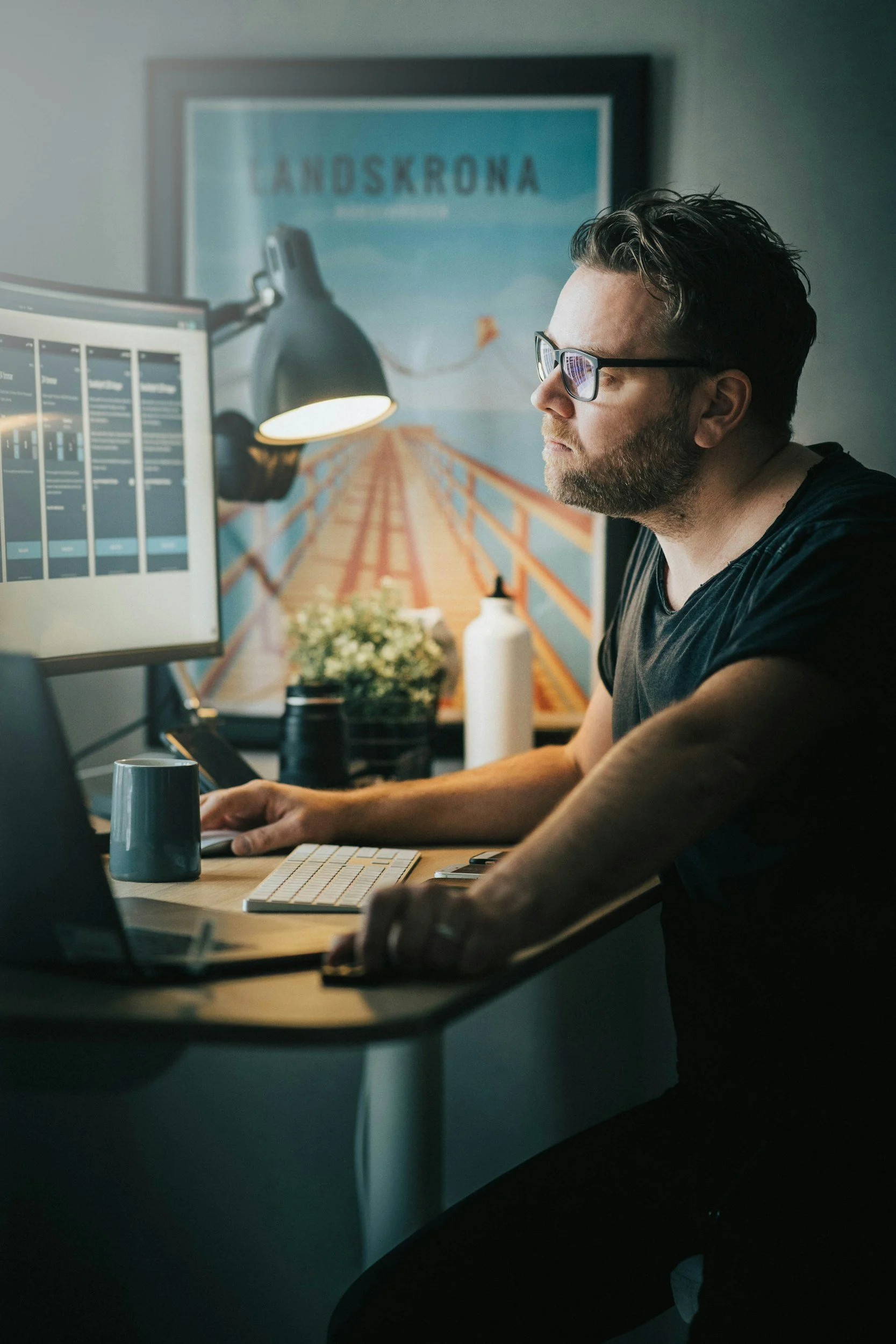 A man with glasses sitting at a desk working on a computer, with posters and objects on the desk.