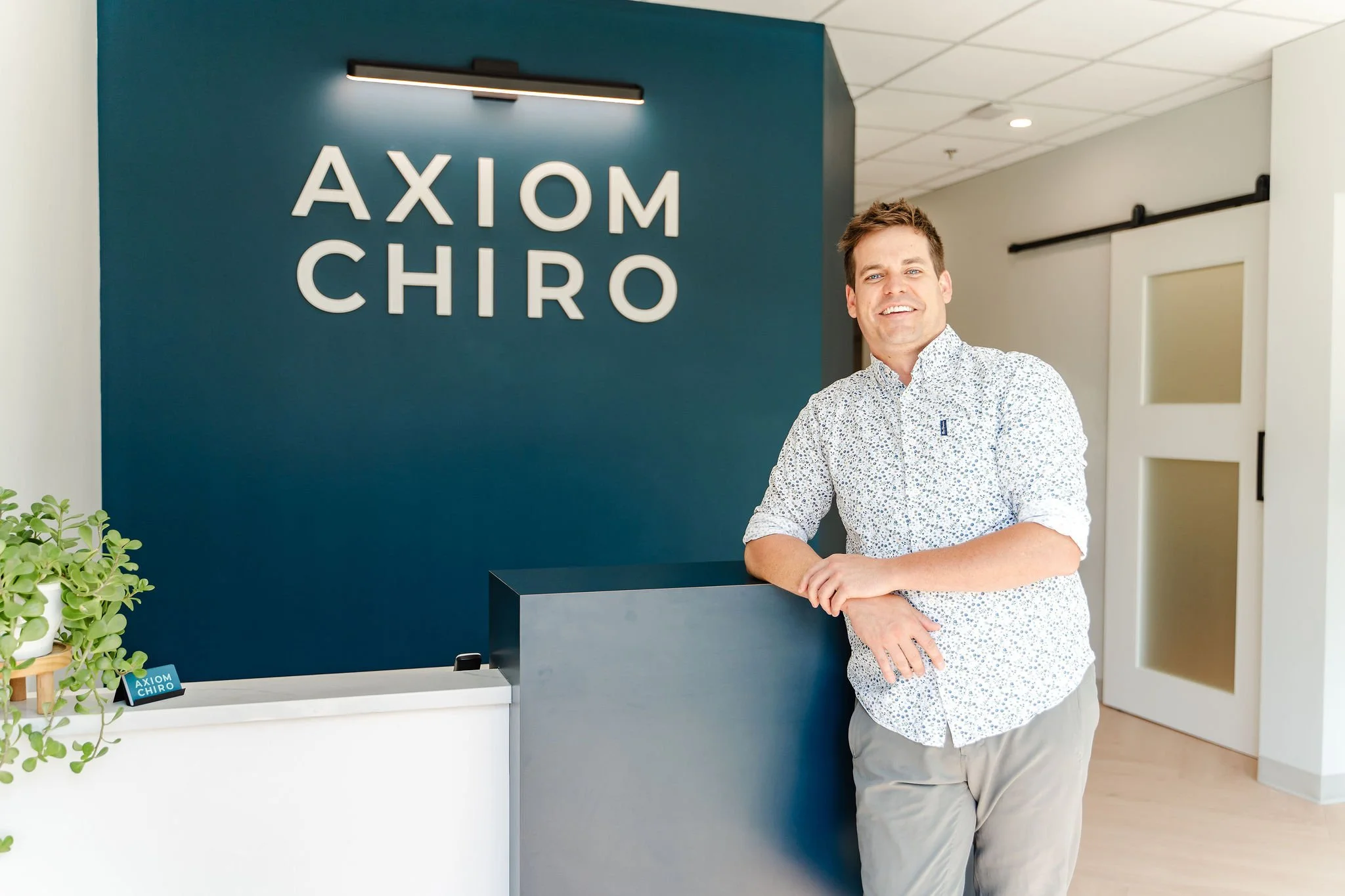 A Calgary chiropractor standing at the reception desk in a modern chiropractor clinic with a sign that reads 'Axiom Chiro' on a blue wall behind him. He is smiling, wearing a patterned shirt, and leaning on the desk.