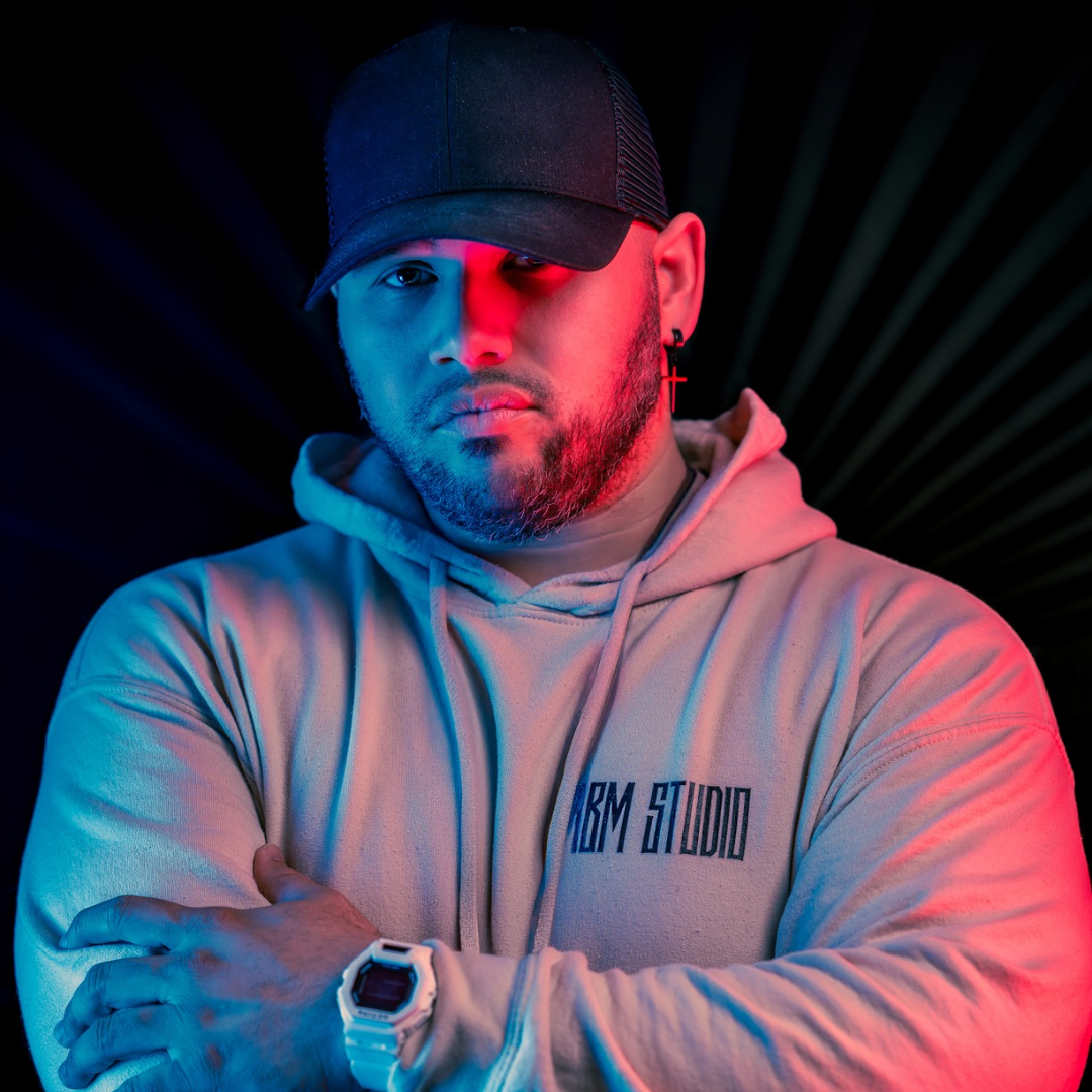 A young man wearing a black cap, hoodie, and white watch, with a cross-shaped earring, standing with his arms crossed against a dark background with colorful lighting. Joseph Correa, Media Manager and Drummer for Philip Mahoney. Filipino Kenny G.