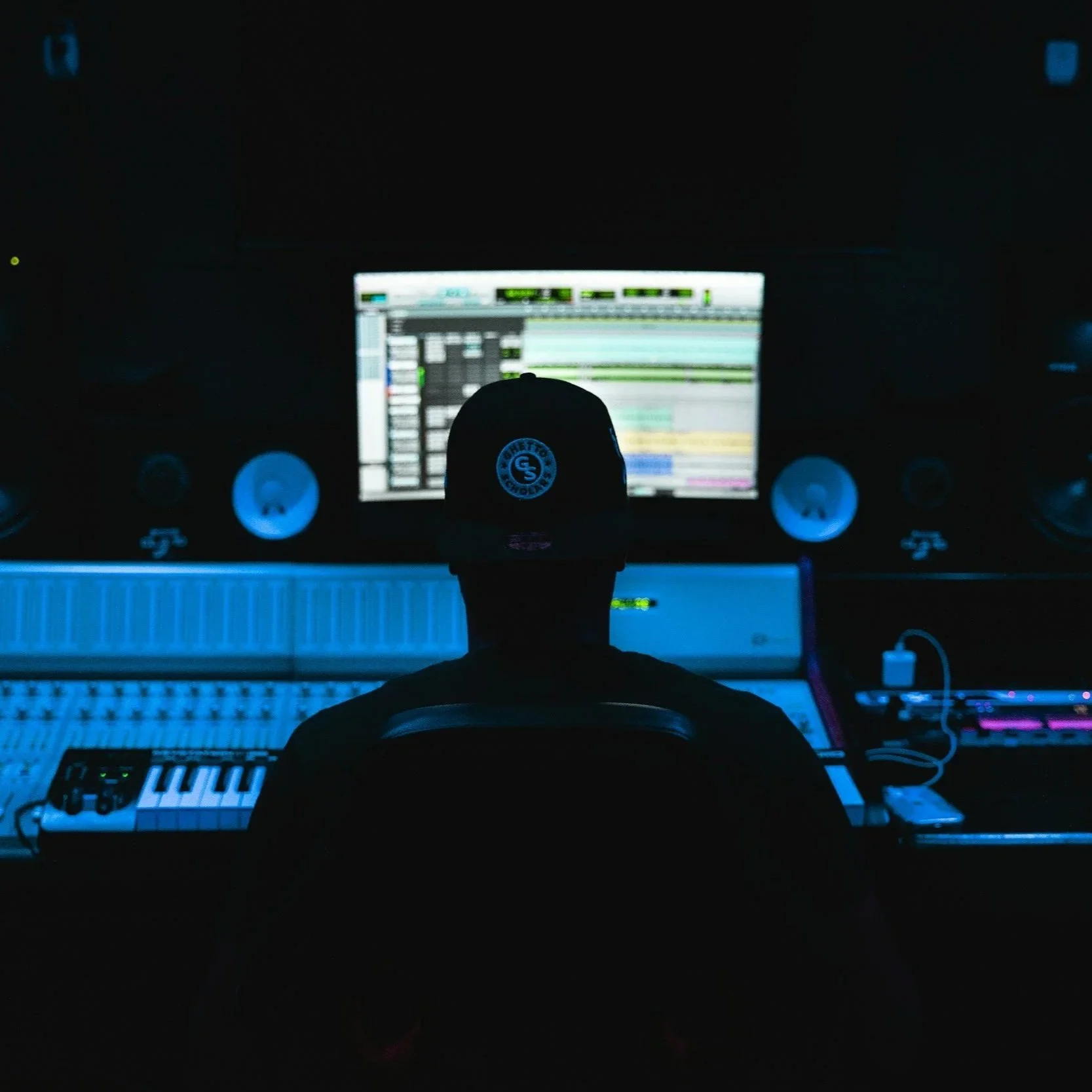 A person in a dark room working at a music studio with a computer monitor displaying digital audio workstation software, surrounded by studio equipment and speakers.
