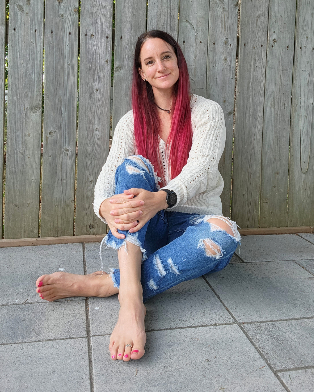 A young woman with long pink hair, wearing a white sweater and ripped jeans, sitting on grey tiled ground with a wooden fence in the background.