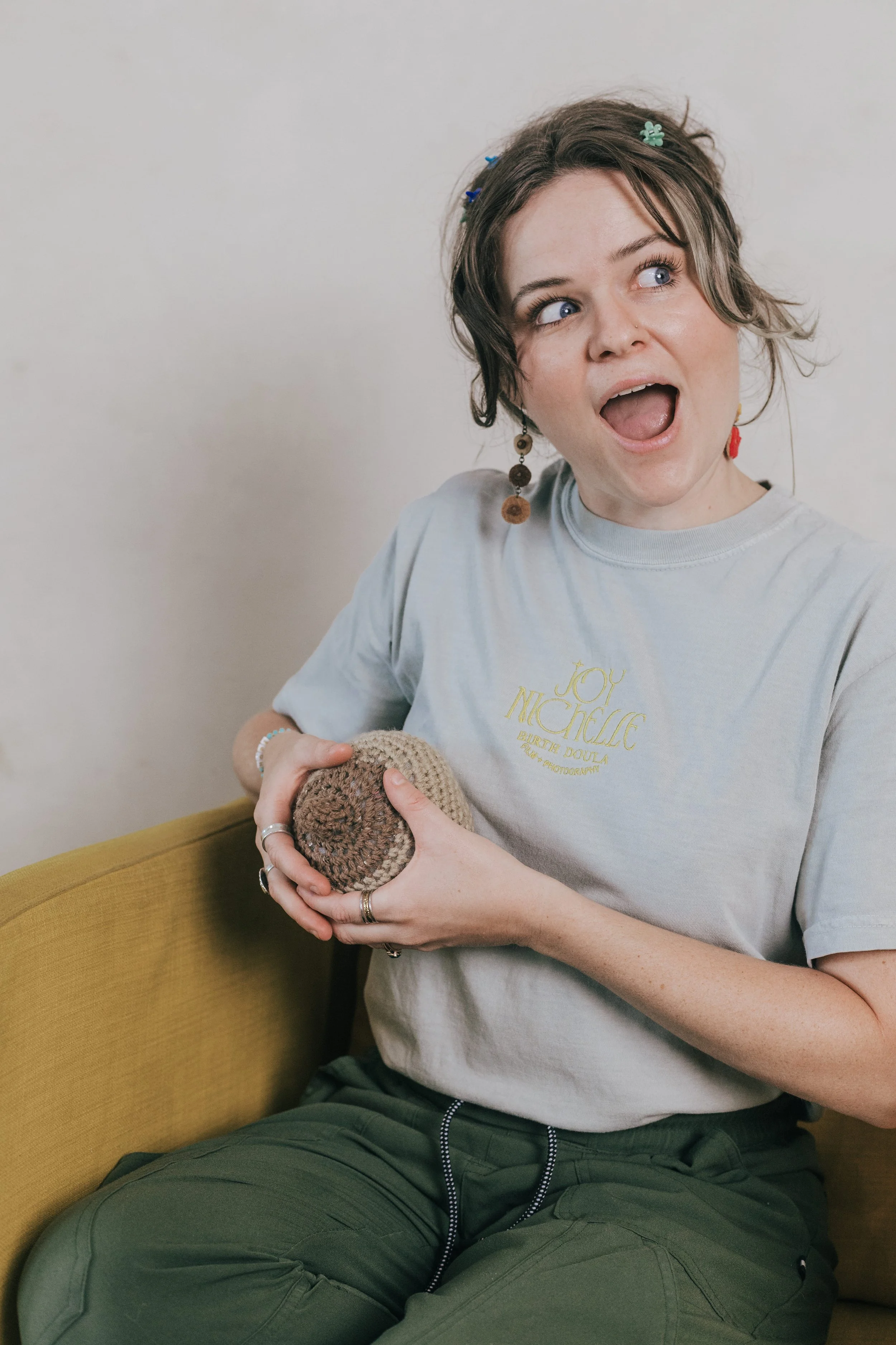 A young woman with short, wavy brown hair adorned with colorful flowers, wearing a light gray T-shirt with gold lettering and dark green pants, sitting on a yellow sofa, holding a small crocheted item, with her mouth open and eyes looking upwards.