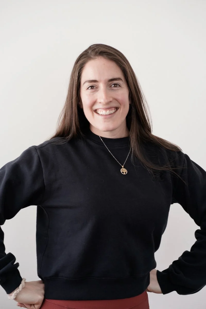 A young woman with long brown hair smiling, wearing a black sweatshirt, a necklace, and a beige scrunchie on her wrist, standing against a plain white background.