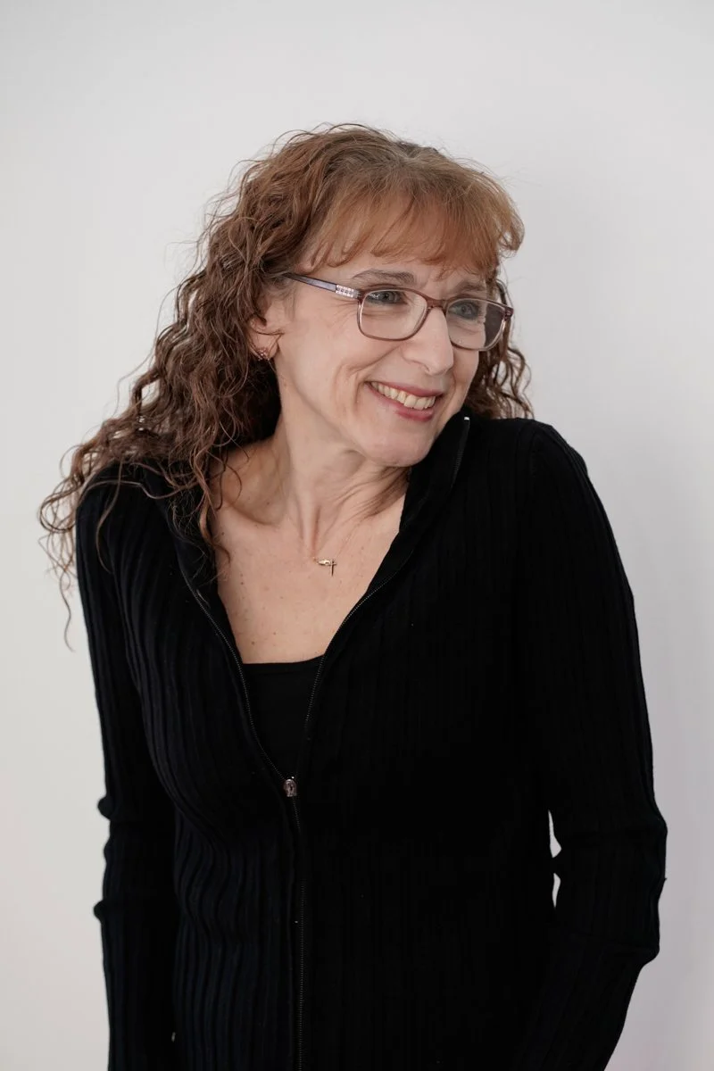 A woman with curly red hair, glasses, and a black zip-up top smiling against a plain white background.