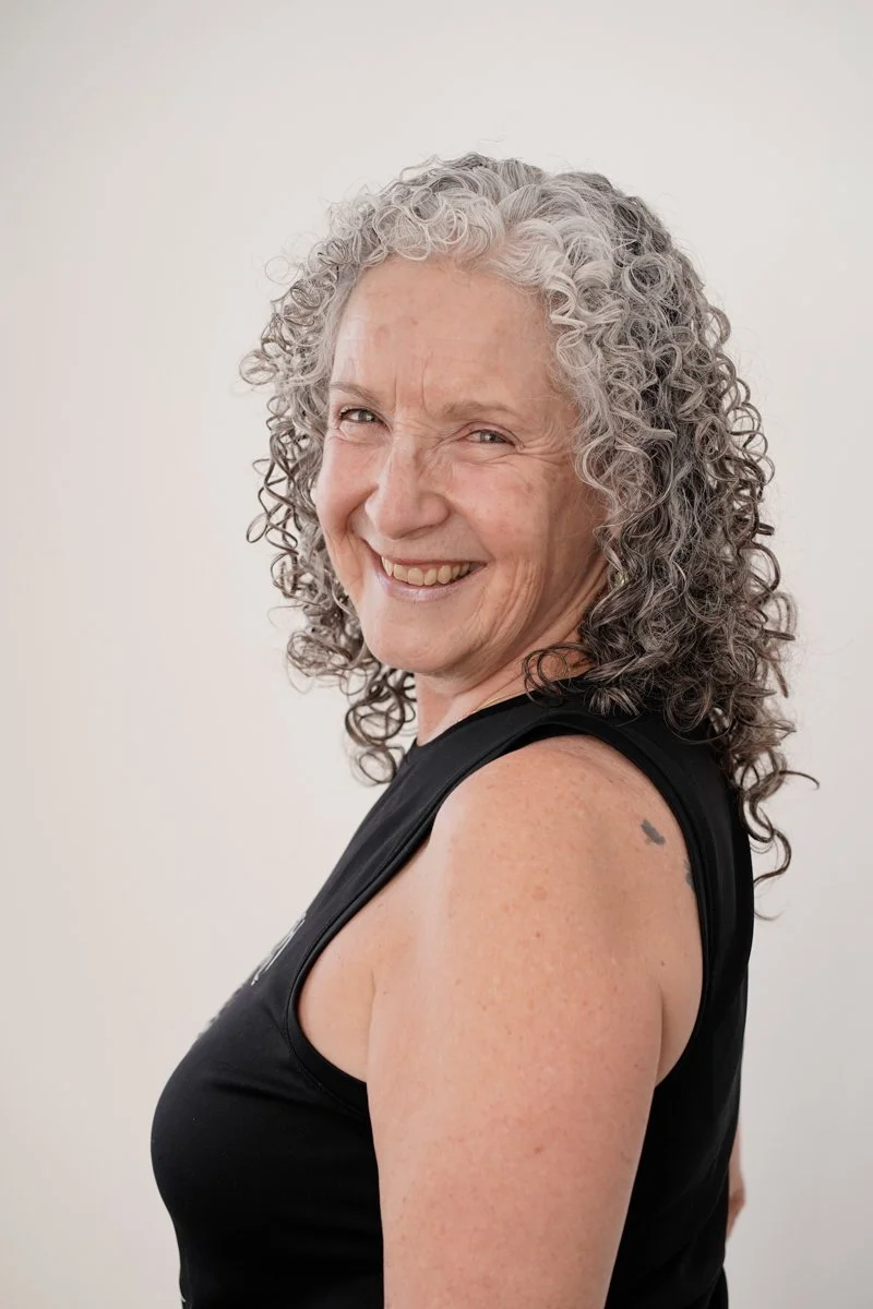 A woman with gray curly hair smiling, wearing a black sleeveless top, standing against a plain white background.