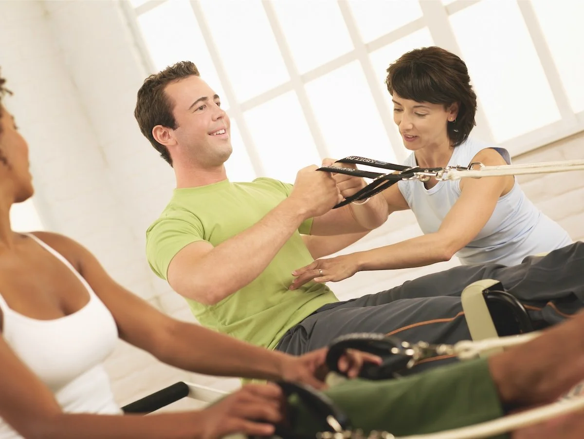 People participating in a Pilates class with suspension trainers, including a man in a green shirt smiling and a woman assisting him, with others in the background.