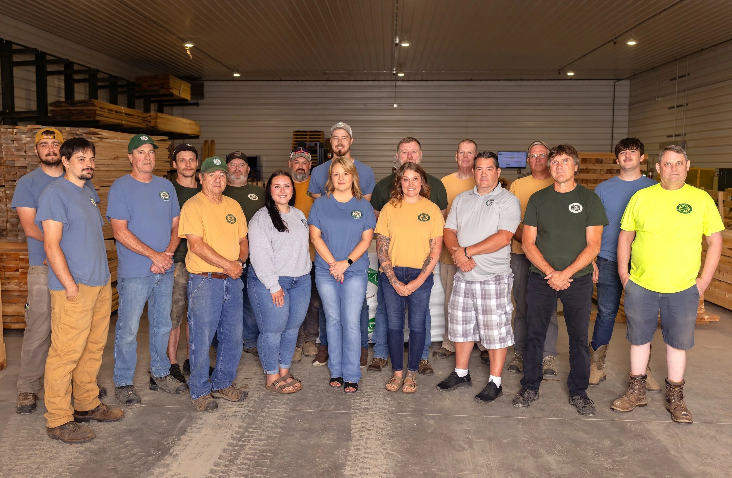 Group of men and women standing inside a woodworking shop, some wearing work shirts, with stacks of wood in the background.