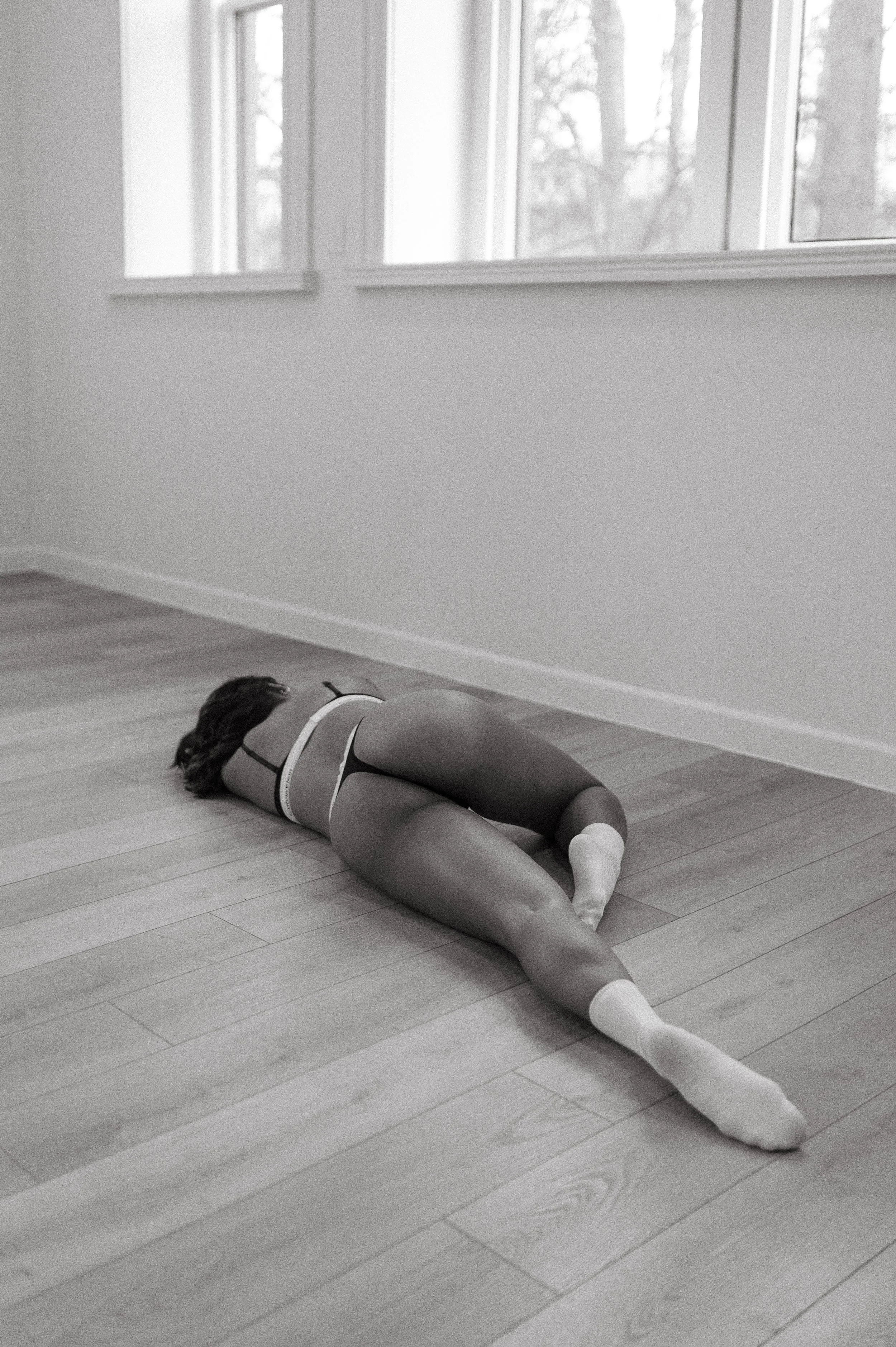 A woman lying on her side on a wooden floor, facing away, wearing underwear and socks, next to a wall and large windows.