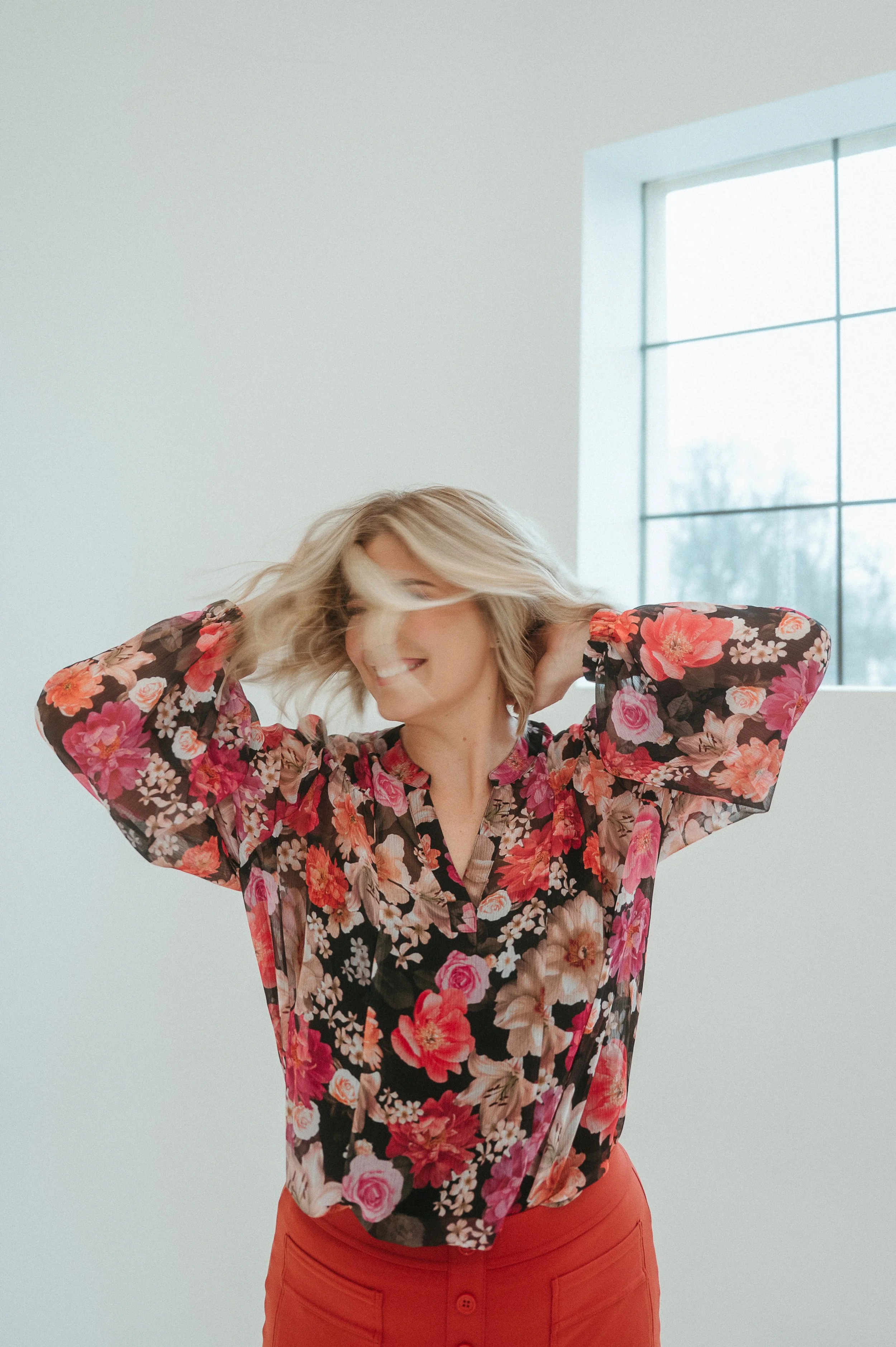 Branding photo of a woman with blonde hair smiling and shaking her head to the side in front of a large window, wearing a floral blouse with red, pink, and white flowers and red pants.