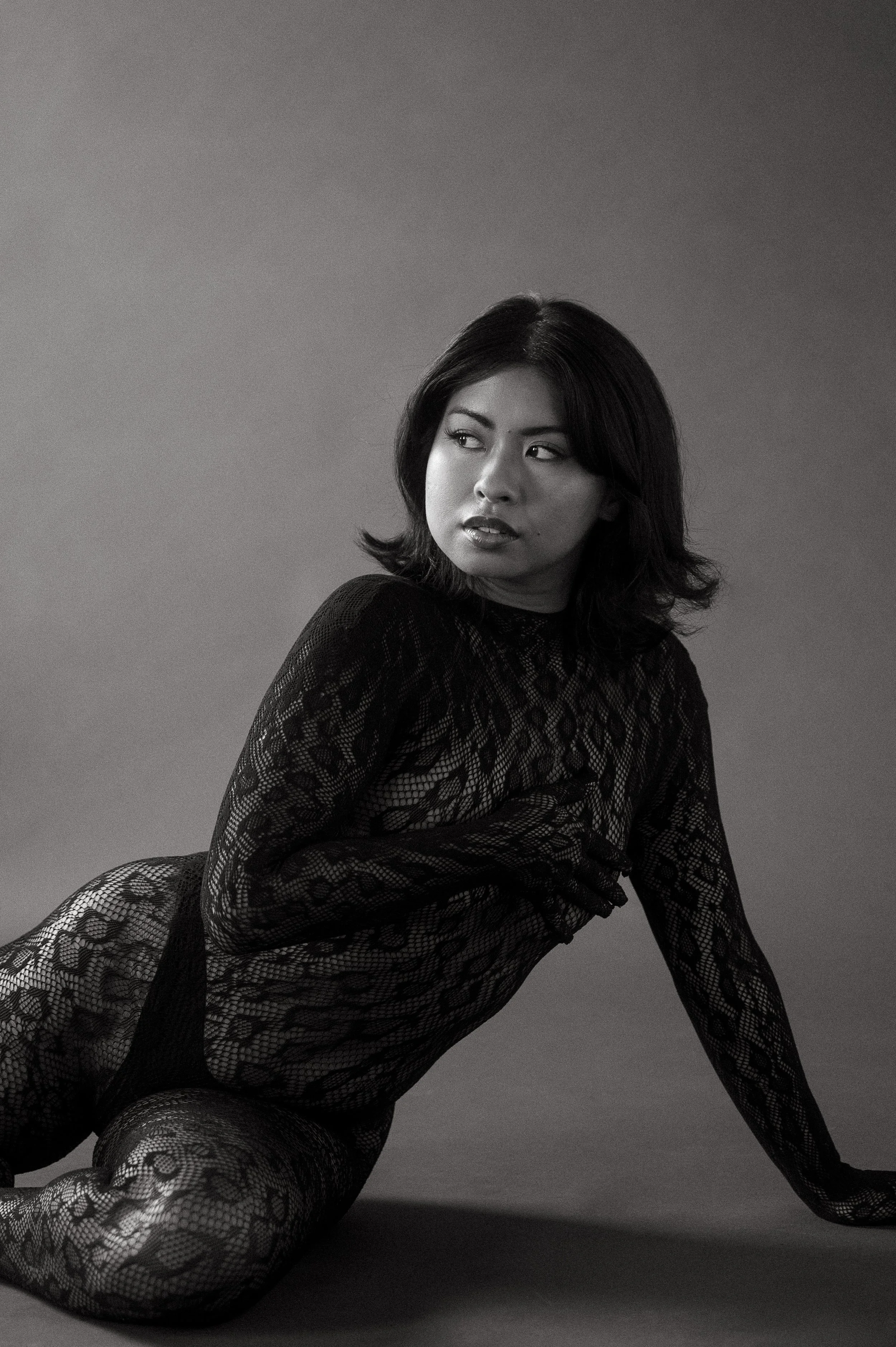Black and white portrait photo of a woman with dark hair, wearing a lace bodysuit, posing on the ground with her hand supporting her body, looking to the side against a plain background.