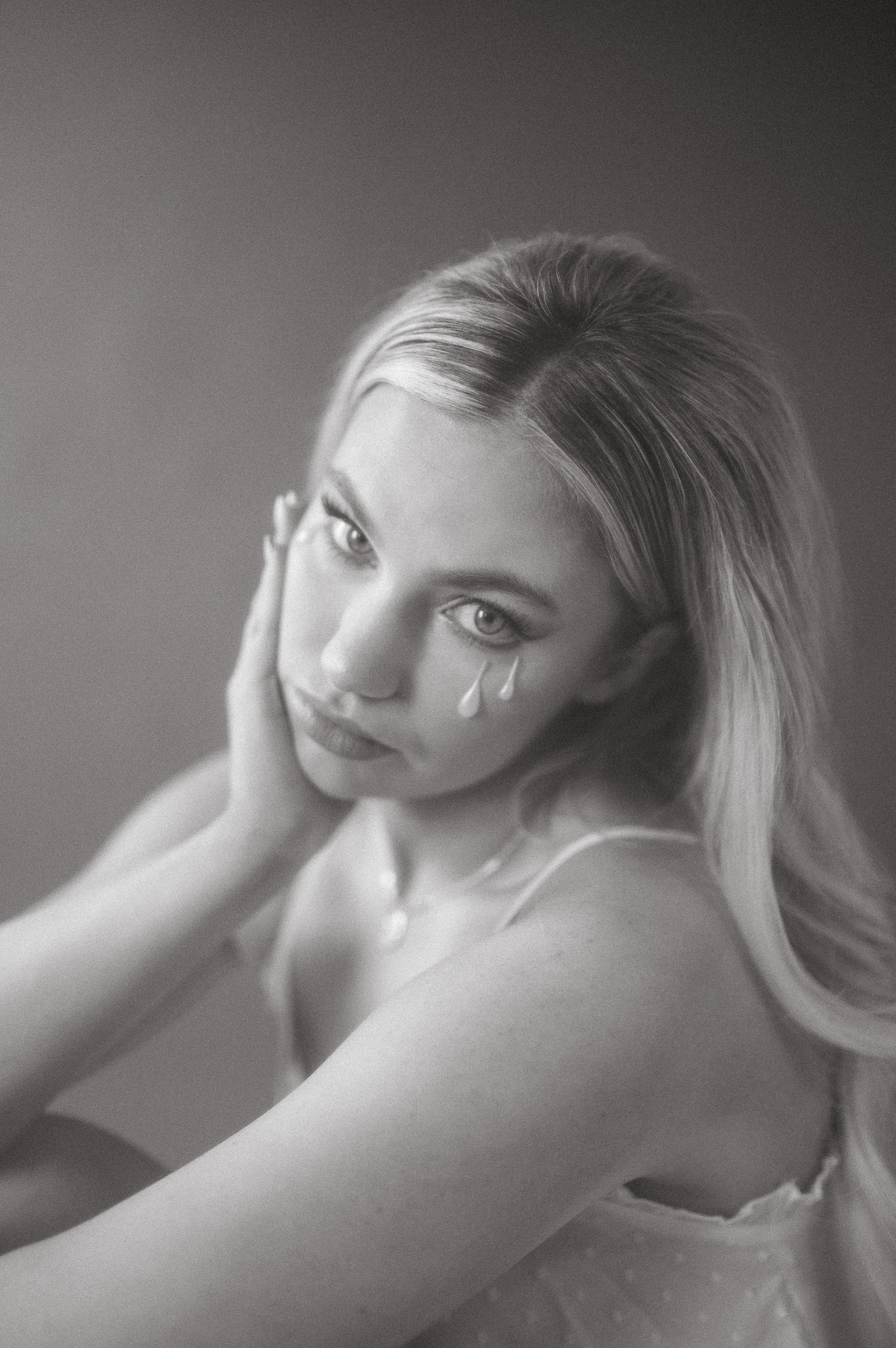 Black and white portrait of a young woman with light-colored hair, looking at the camera with a serious expression, resting her face on her hand, with tears illustrated on her cheek.
