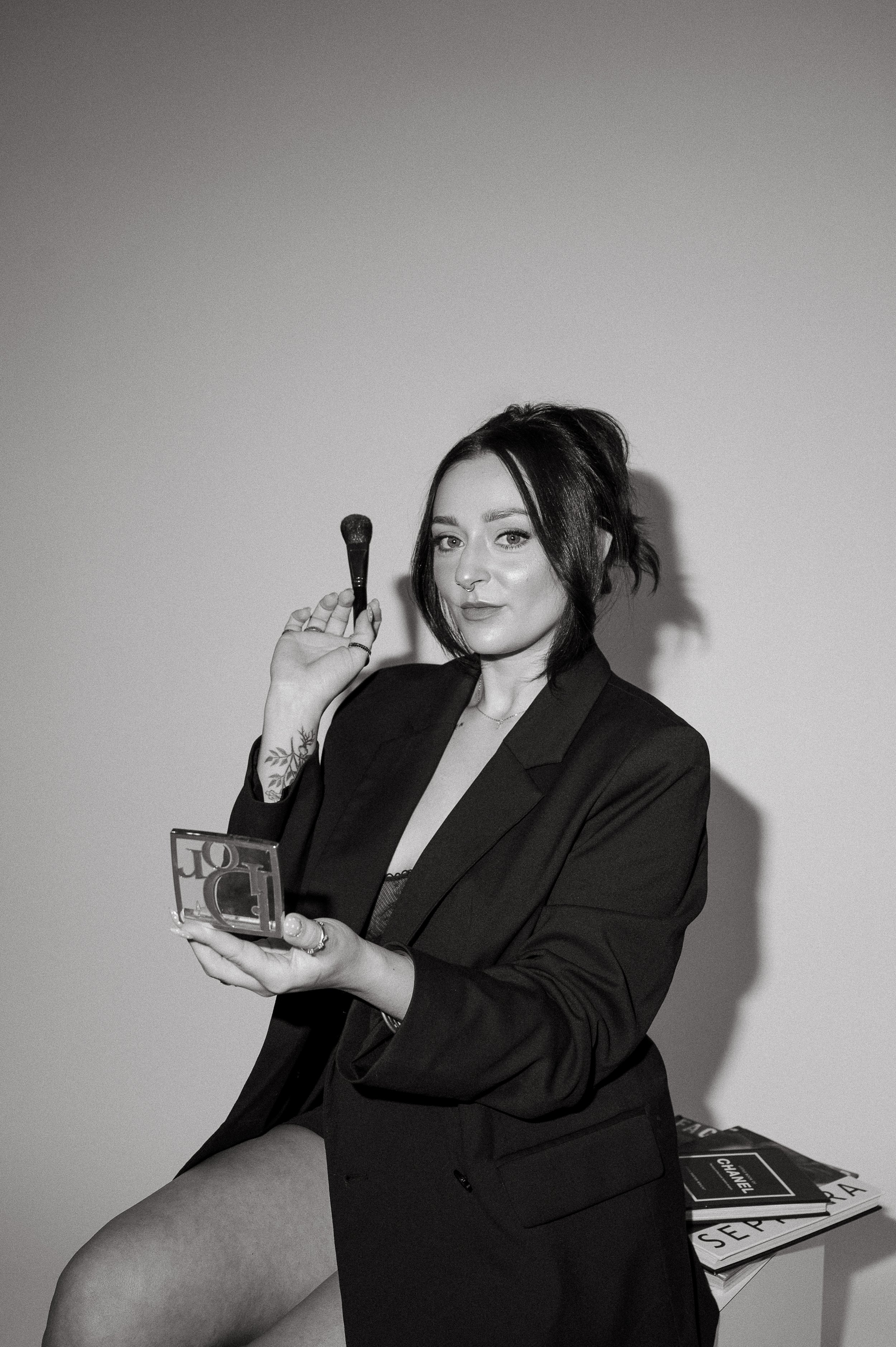 Black and white branding photo of a woman with dark hair in a messy bun, wearing a blazer, holding a makeup brush in one hand and a compact mirror in the other, sitting on a table with magazines.