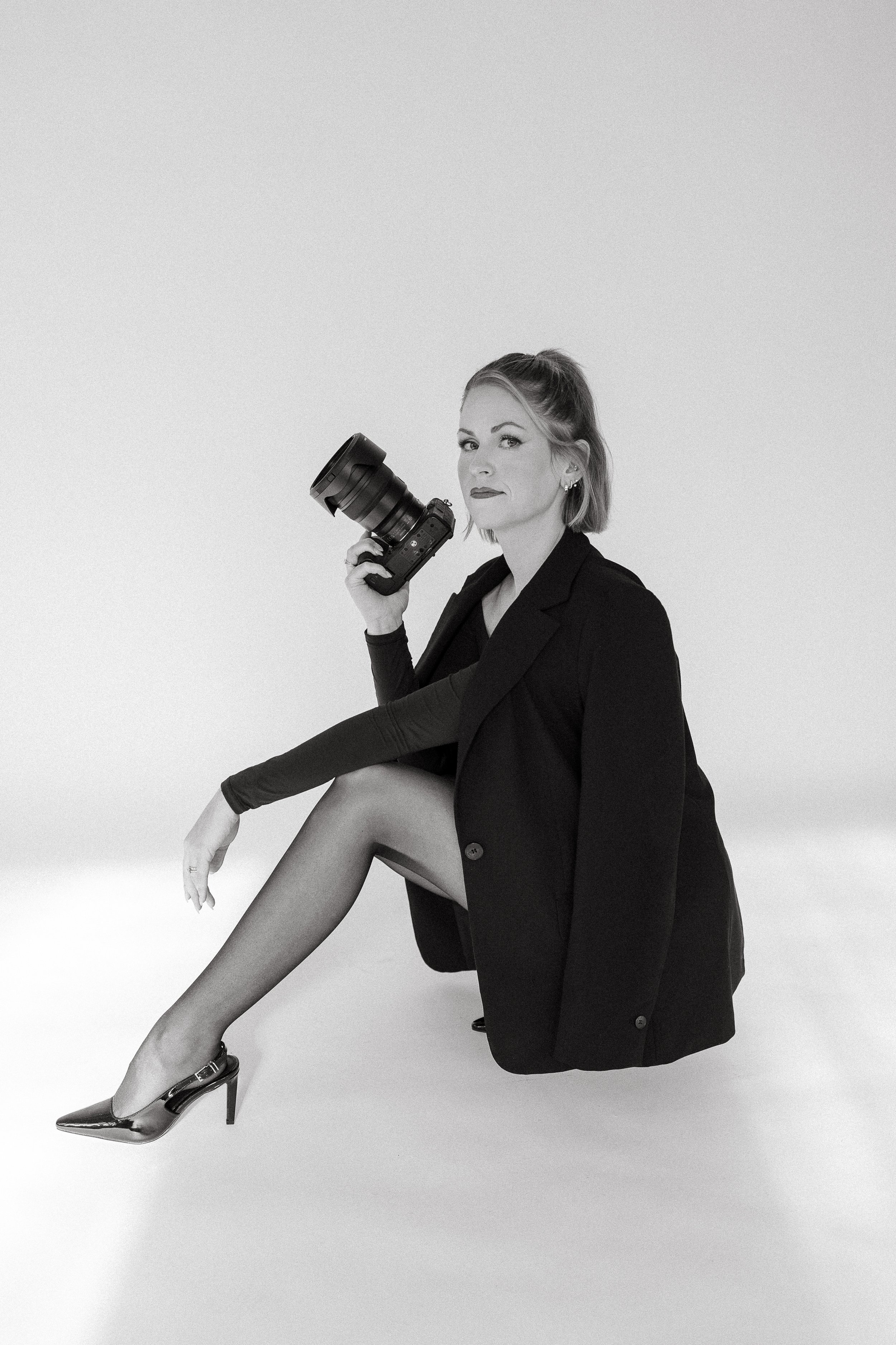 Black and white photo of a woman sitting with one leg crossed over the other, wearing high heels, a blazer, sheer stockings, and holding a professional Nikon brand camera, on a plain white background.