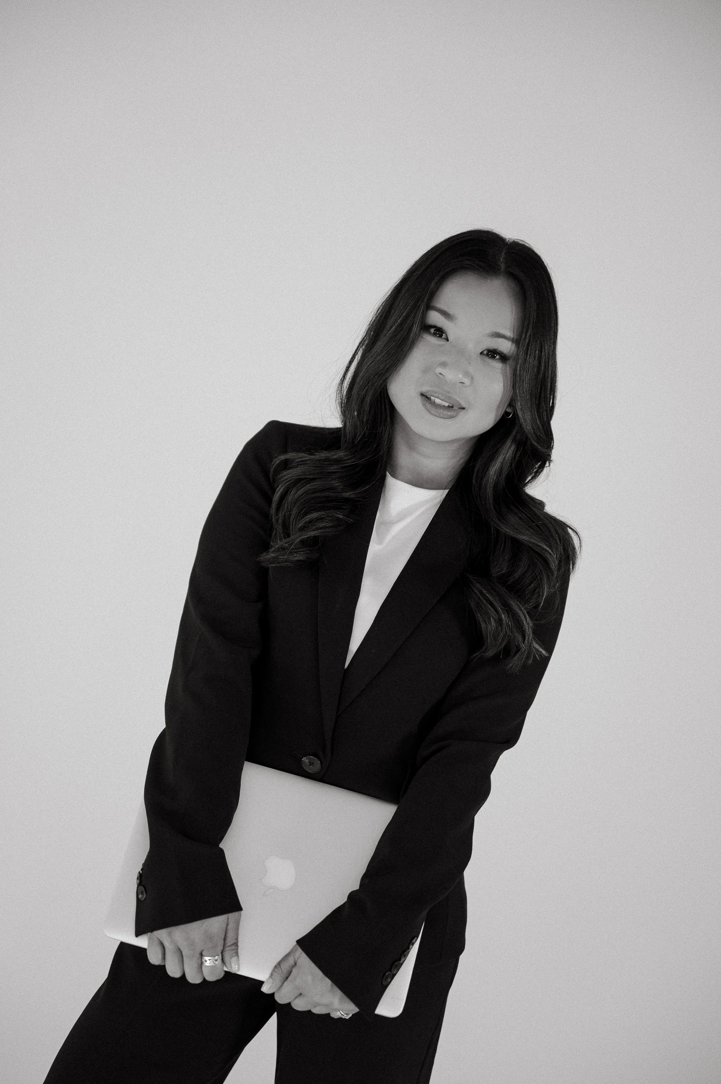 Black and white branding photo of a woman in a black suit holding a silver MacBook laptop, standing against a plain light background.