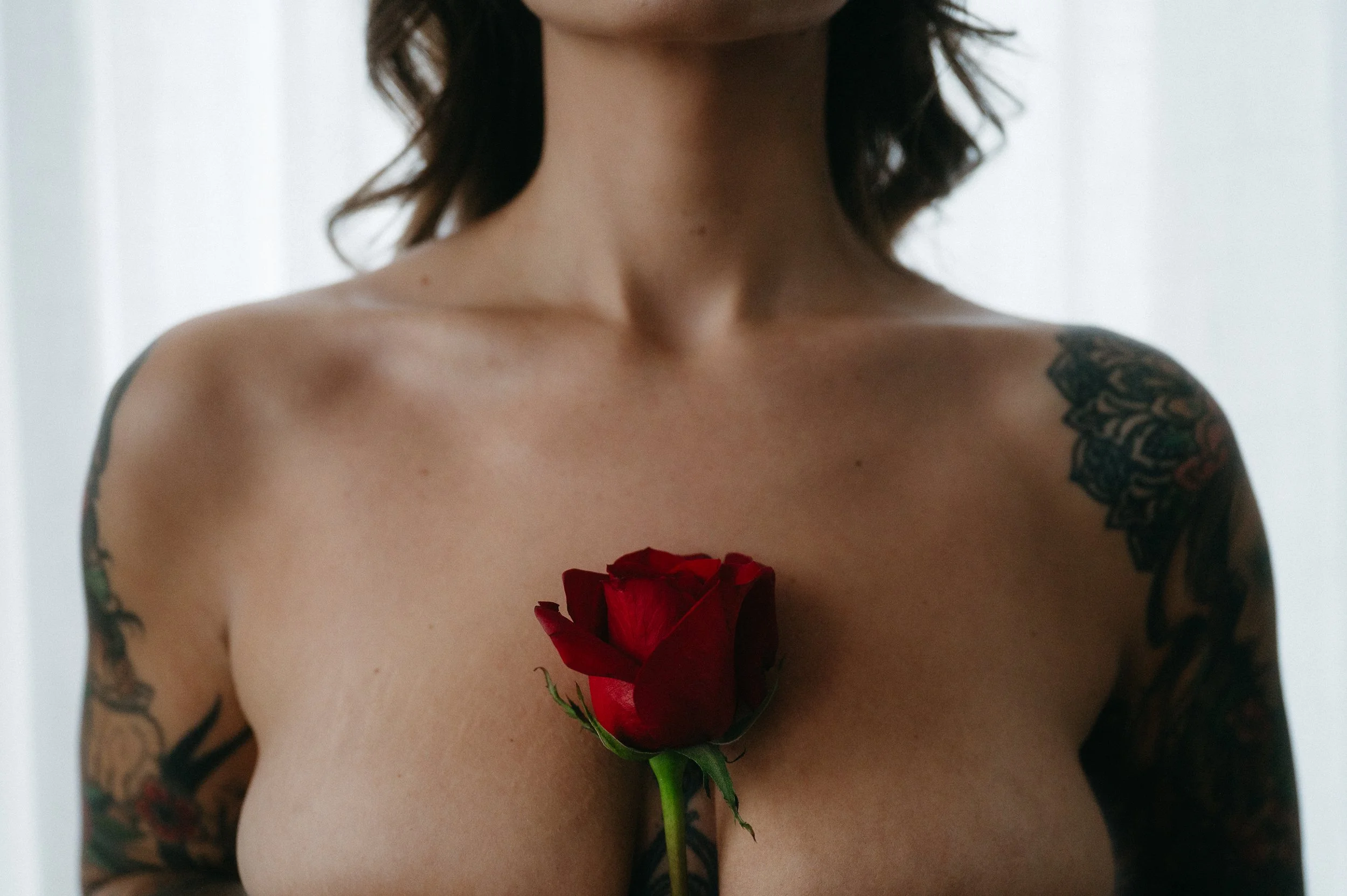 A woman with tattoos on her shoulders holds a red rose in front of her bare chest.