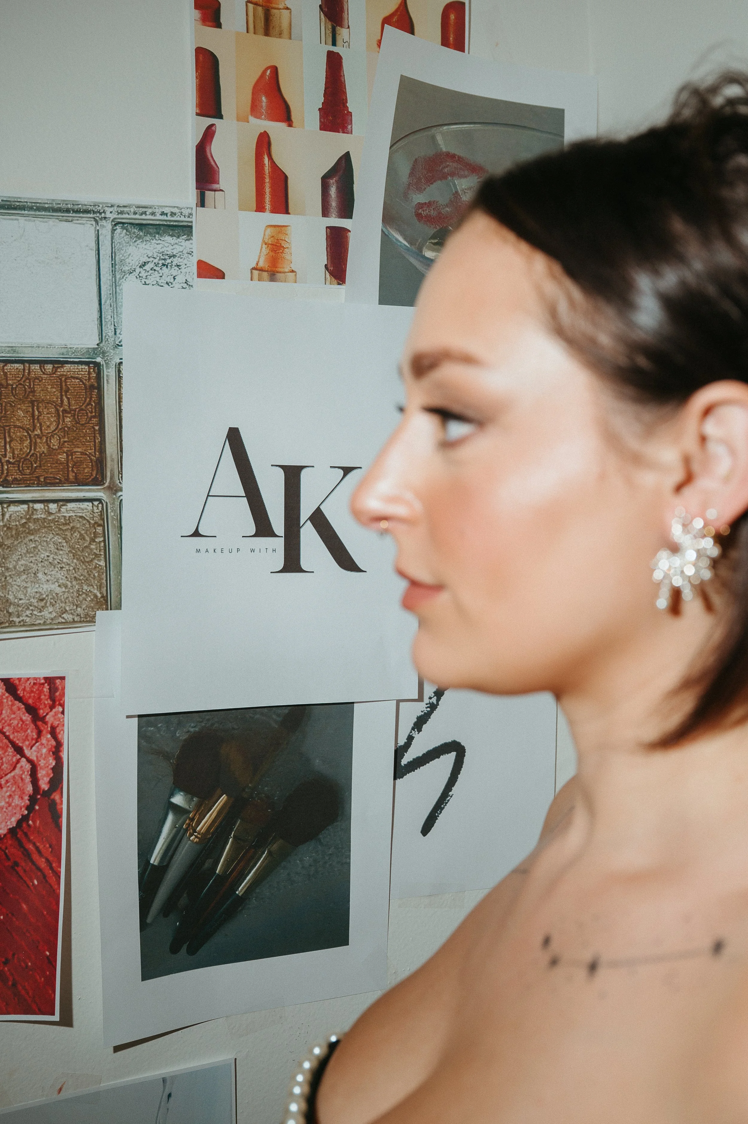 Branding photo of the side profile of a woman with earrings and a nose piercing, standing in front of a wall decorated with makeup and lipstick collage, including a sign that says 'AK'.