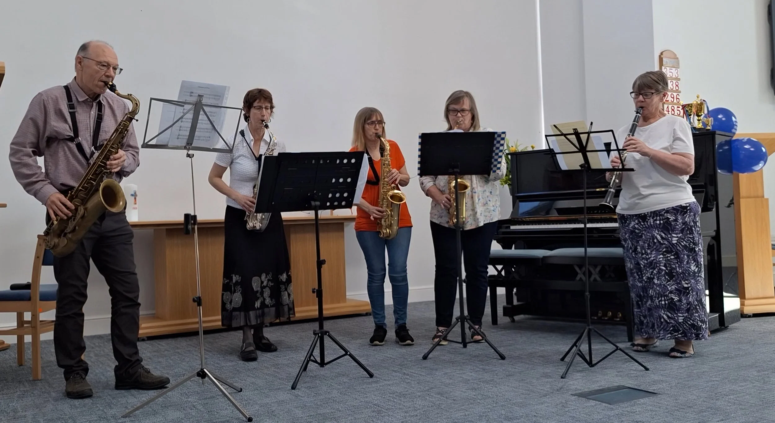 1 tenor, 3 alto sax and one clarinet player stand in front of a piano in a church like setting,  they have music on the stand in front of them.  They are fairly smartly dressed for a concert.