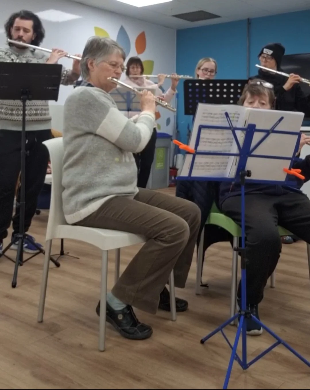 Group of adults sit at music stand in a well lit room, a lady at the front wears her glasses on her head and can barely be seen over her music stand.
