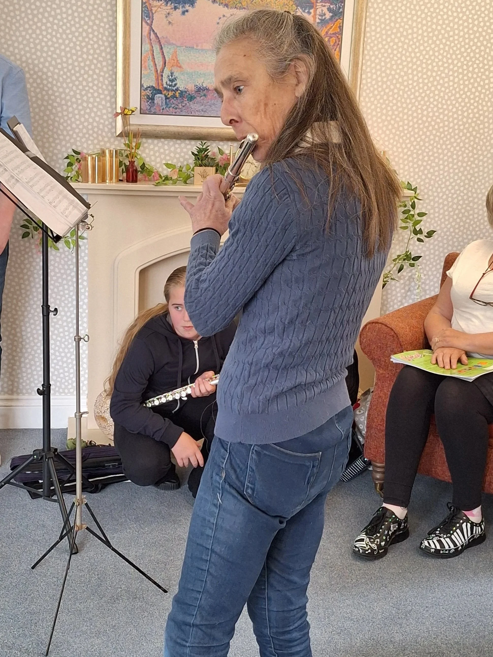 Lady in her 50s playing flute in a care home.  Other music performers sit listening in the backgroun