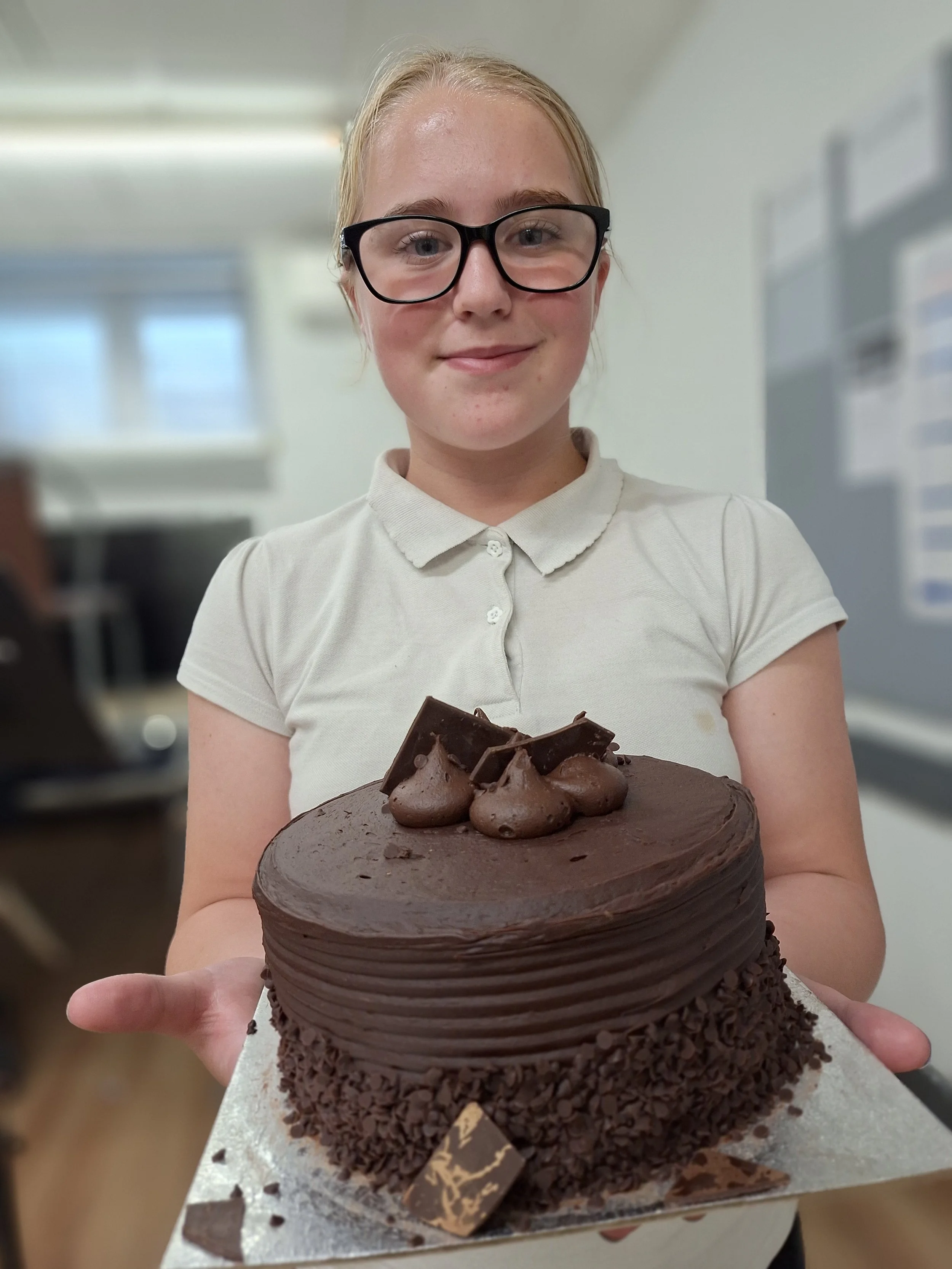 12 year old girl with school polo white t-shirt on and dark rimmed glasses holds huge chocolate cake on a silver stand.  She is an office like room.