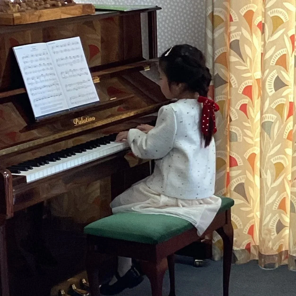 Petite 8 year old girl sits in front of a patterned curtain.  Child sits in light cardigan and skirt and shiny brown piano with music on the stand.