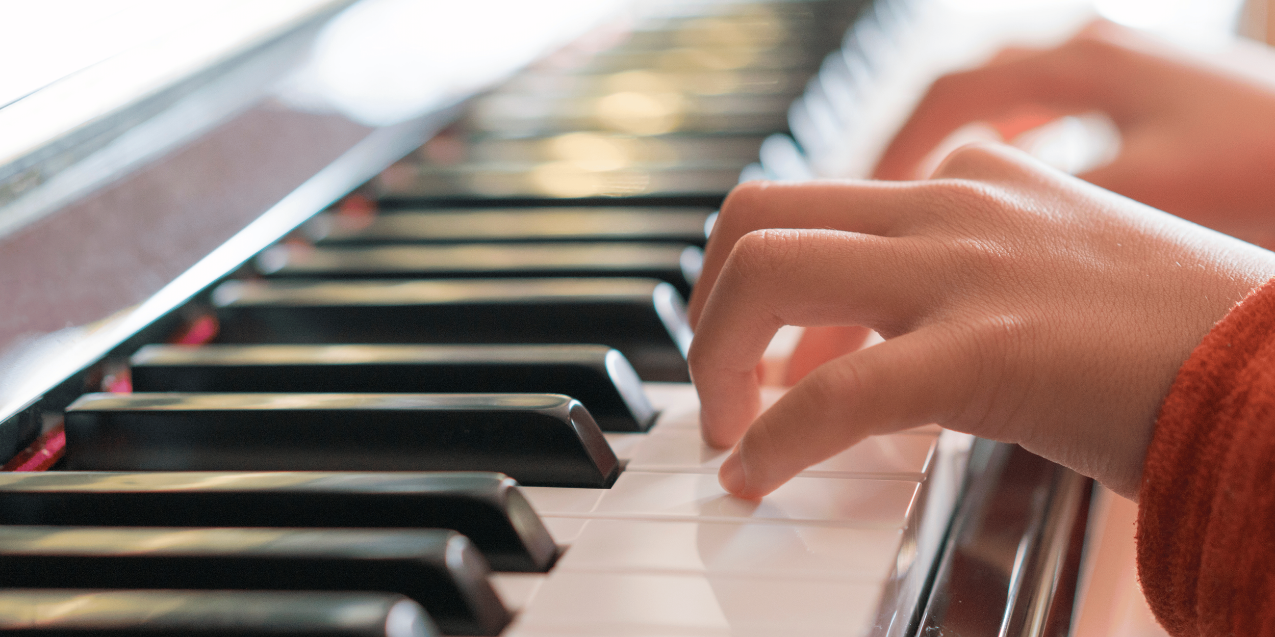 child's hands rest of piano keys, the burnt red cuffs of their jumper are visible