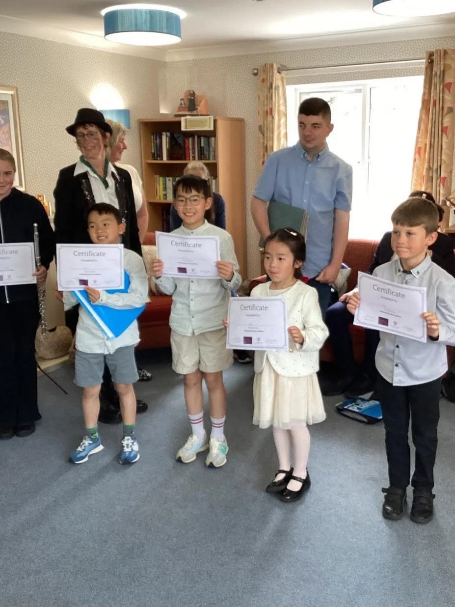 5 children stand holding music certificates in front of other adults who have been performing.  One lady in the background appears to be dressed as a chimney sweep with soot rubbed on her cheeks.  One teen holds a flute.