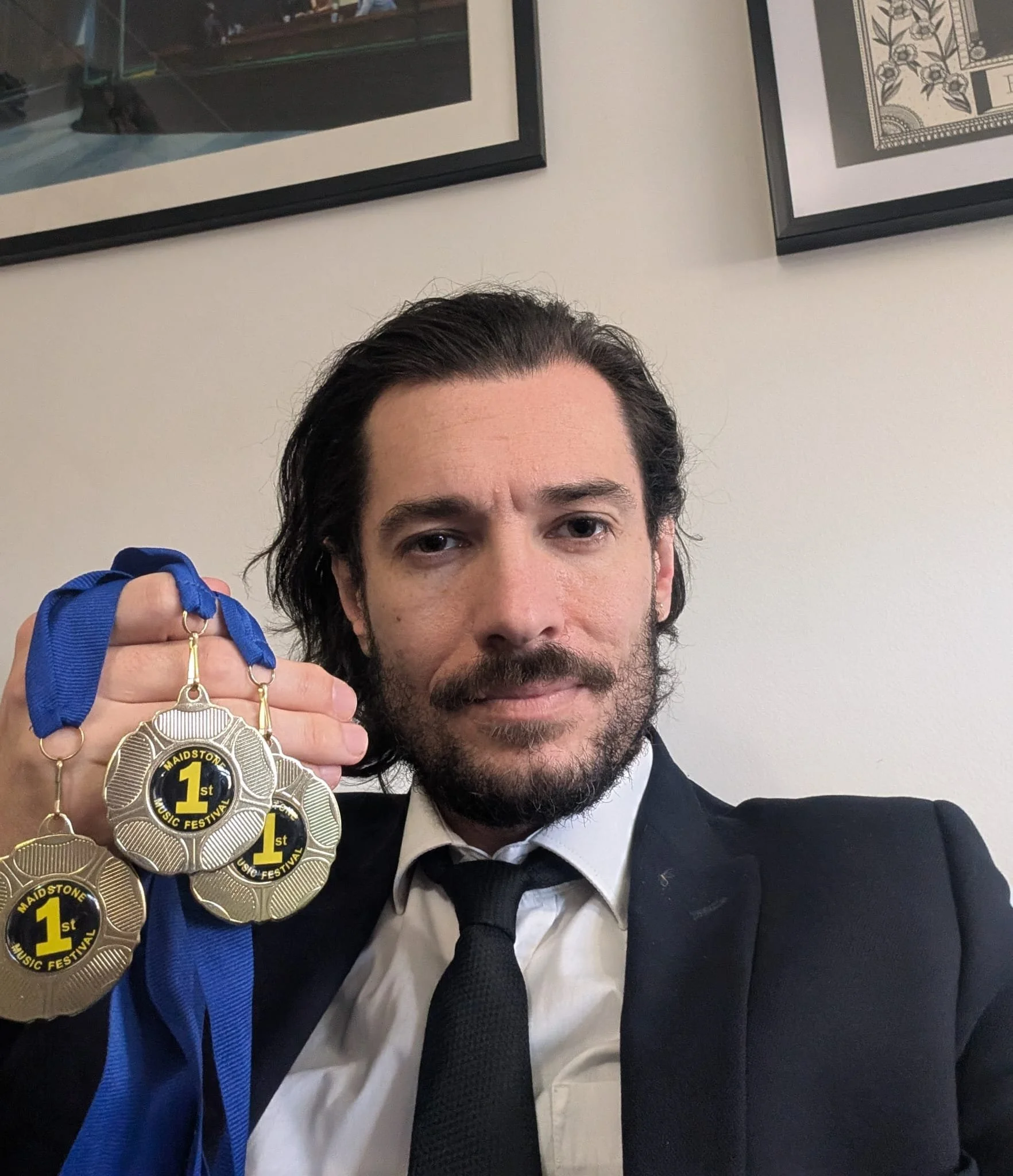 A smart looking man, with suit and black tie, beard, tash and mid length hair neatly swept back sicks proudly holding 3 medals displaying suggest 1st place in 3 music categories.