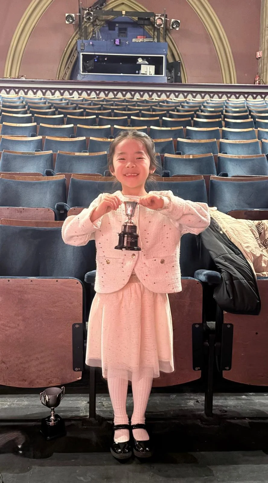 7 year old girl in pretty white skirt, cardigan and black school shoes stands in a theatre in front of rows of empty chairs.  She proudly holds a silver cup.