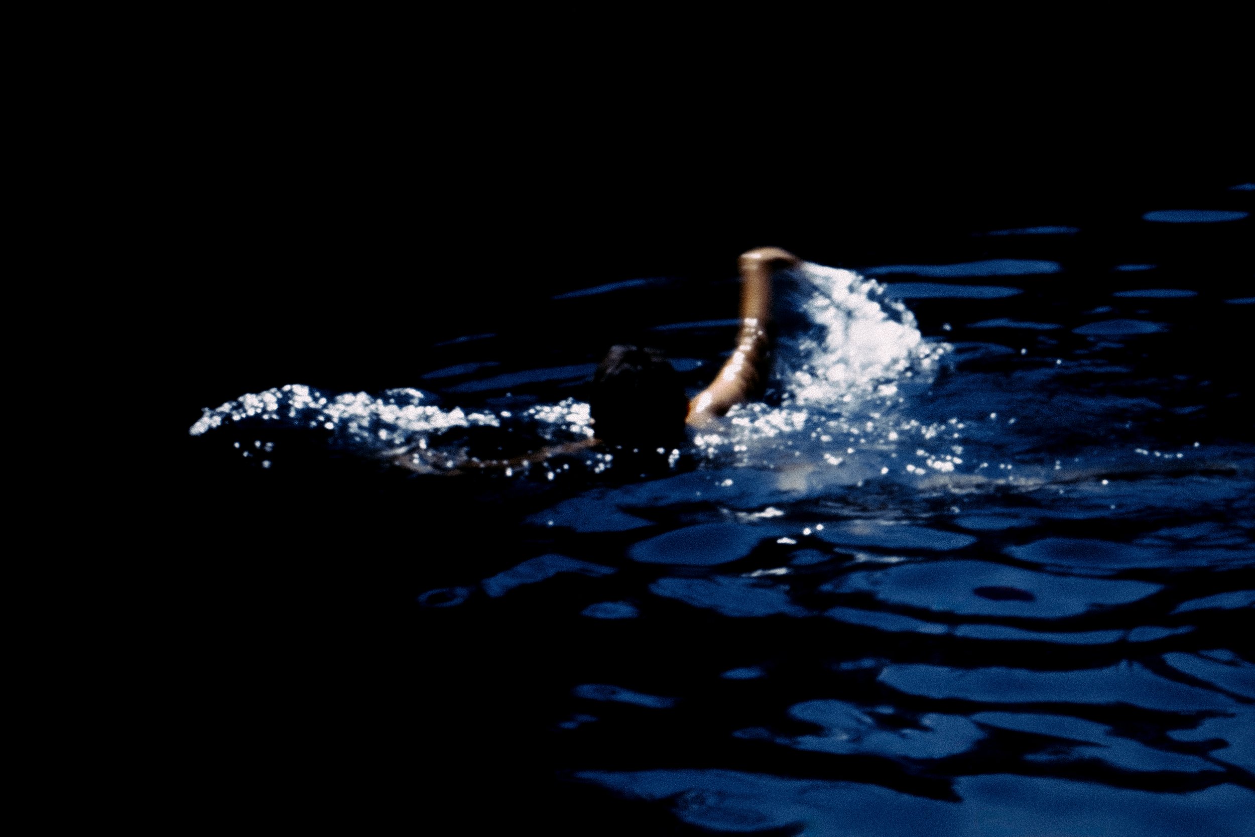 Person swimming in dark water, arm outstretched, creating splashes.