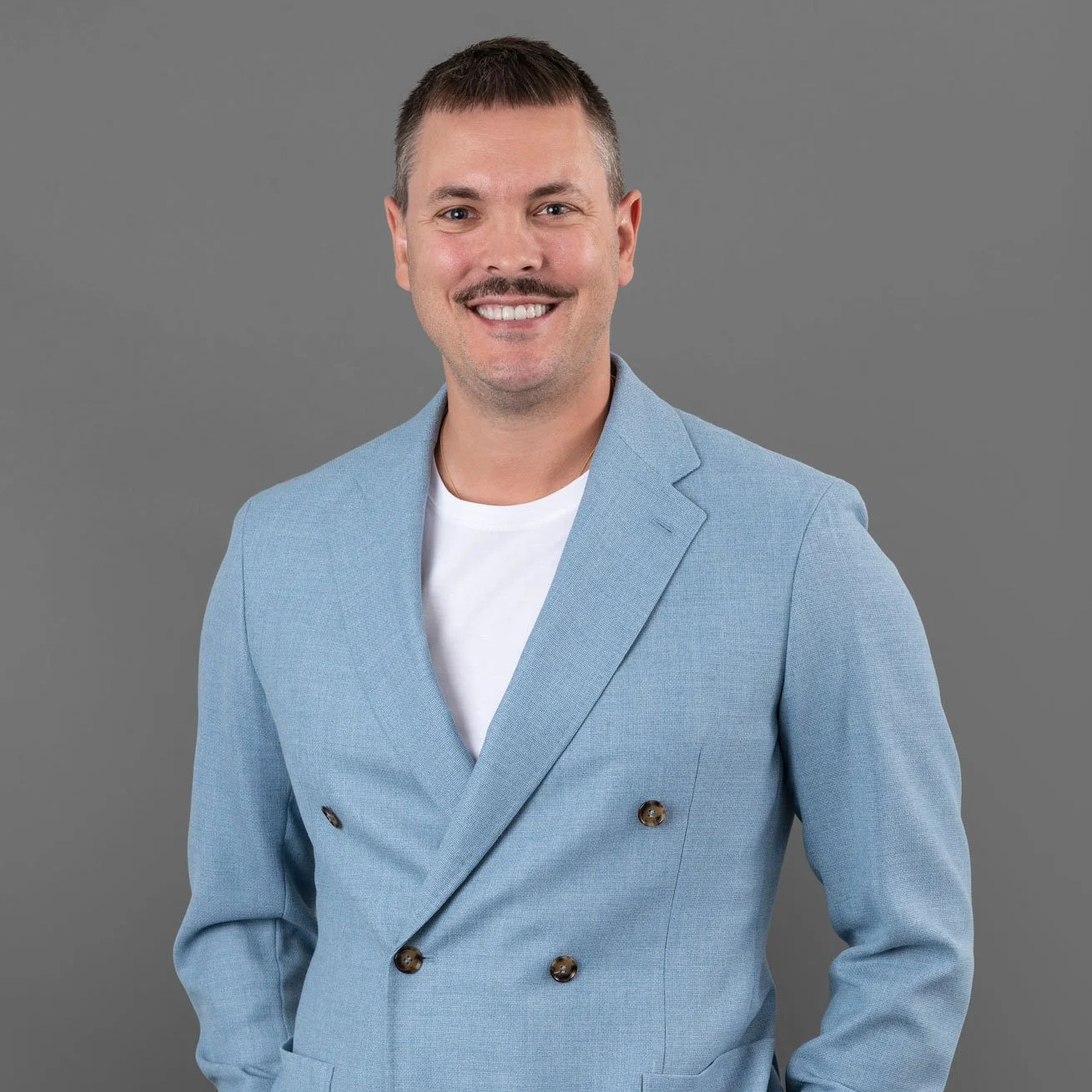 Brian Hemmi trägt einen hellblauen Anzug und lächelt vor hellem grauem Hintergrund.