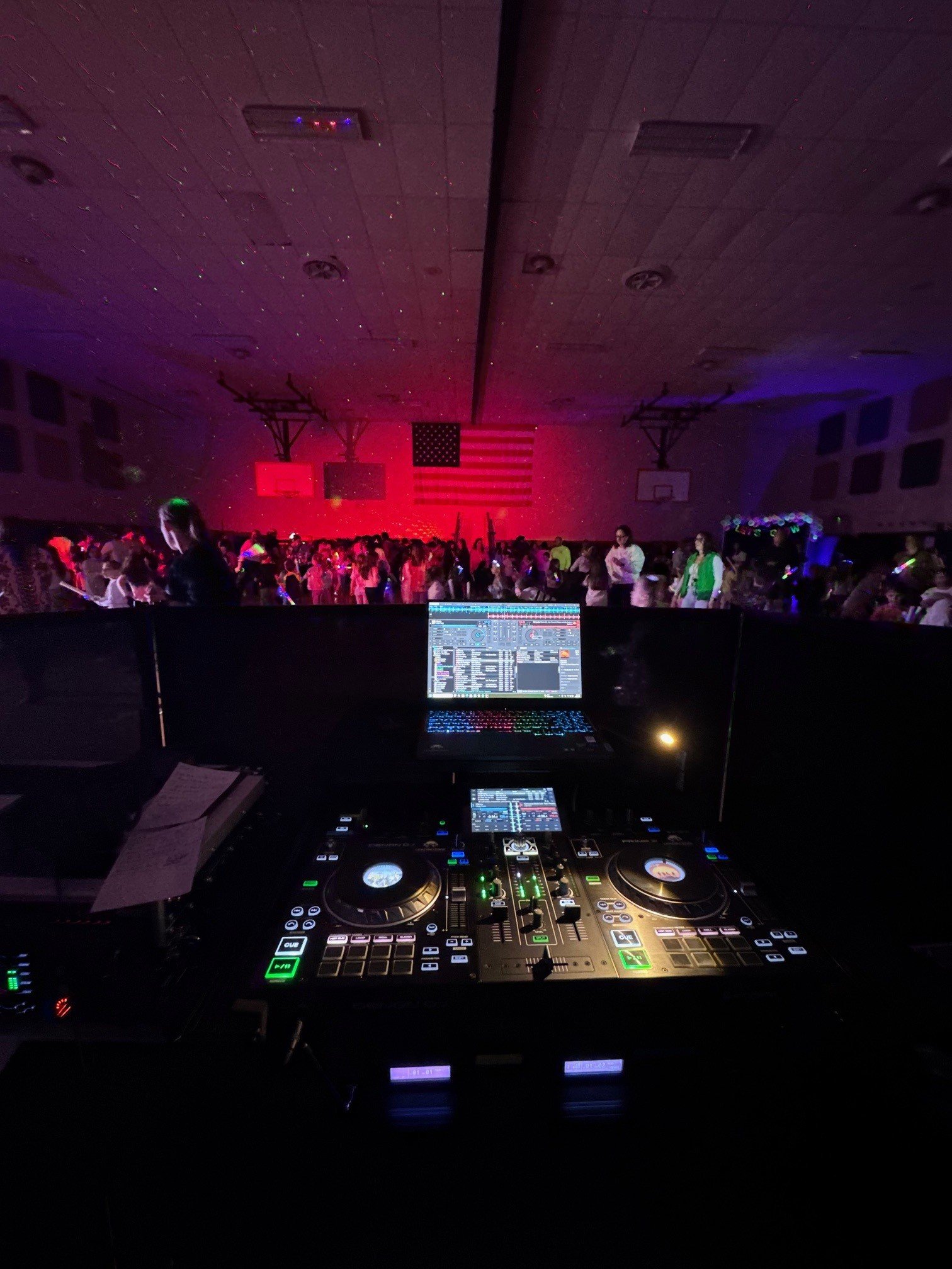 DJ booth with a laptop and sound equipment overlooking a dance floor with people, American flag, and gymnasium ceiling lights at a school dance event.