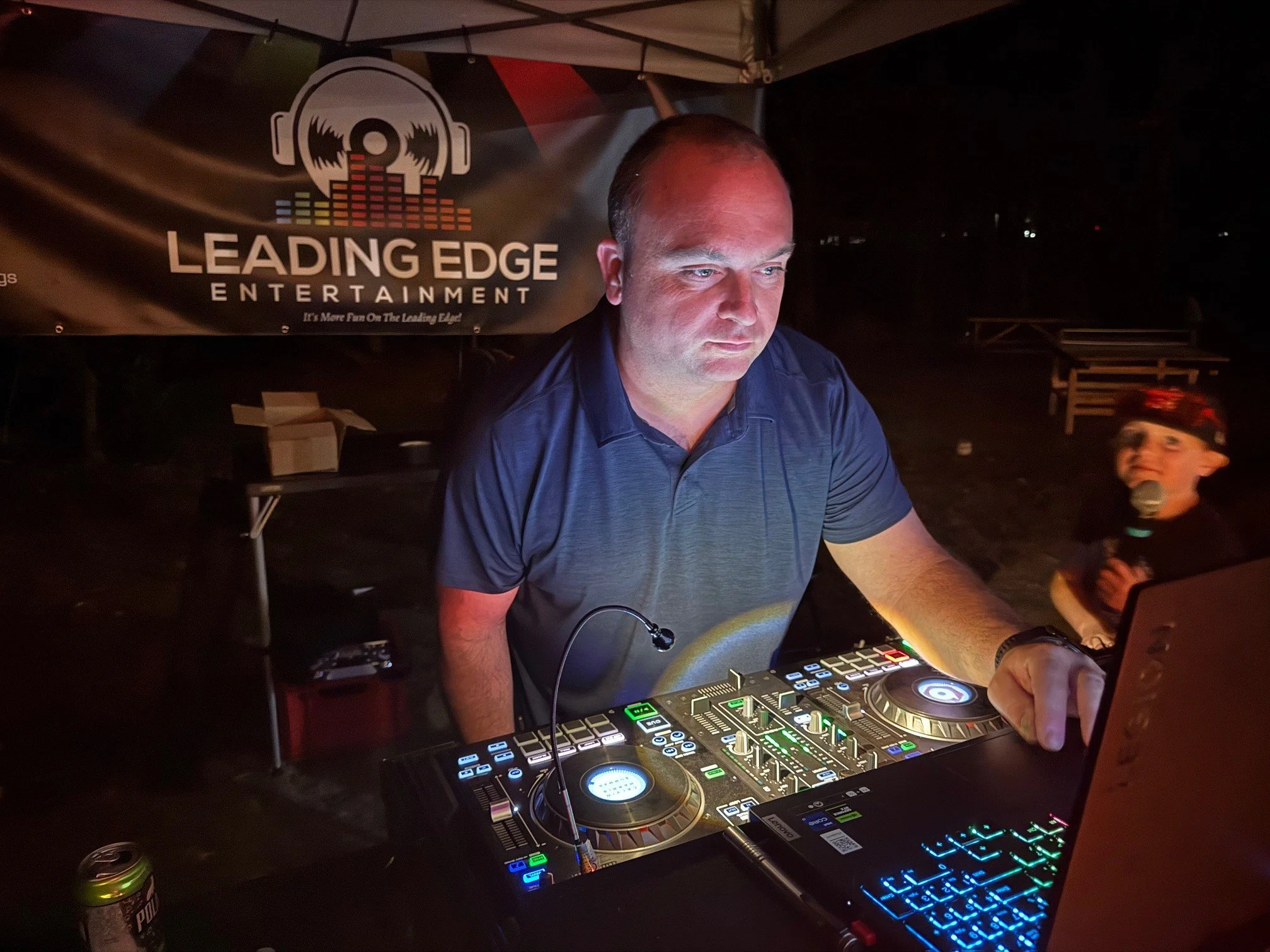 A DJ is performing at an outdoor event during nighttime, wearing a blue shirt and standing behind a DJ controller with colorful lights. There is a young boy wearing a cap and holding a microphone in the background. A banner with the logo and name "Leading Edge Entertainment" is visible behind him.