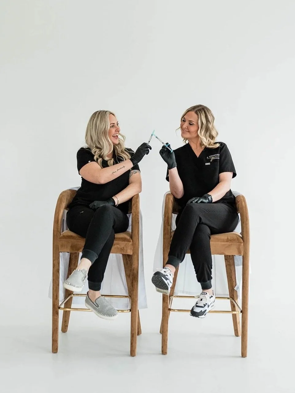 Two women in black medical scrubs sitting on wooden chairs, smiling, exchanging syringes, against a plain white background.