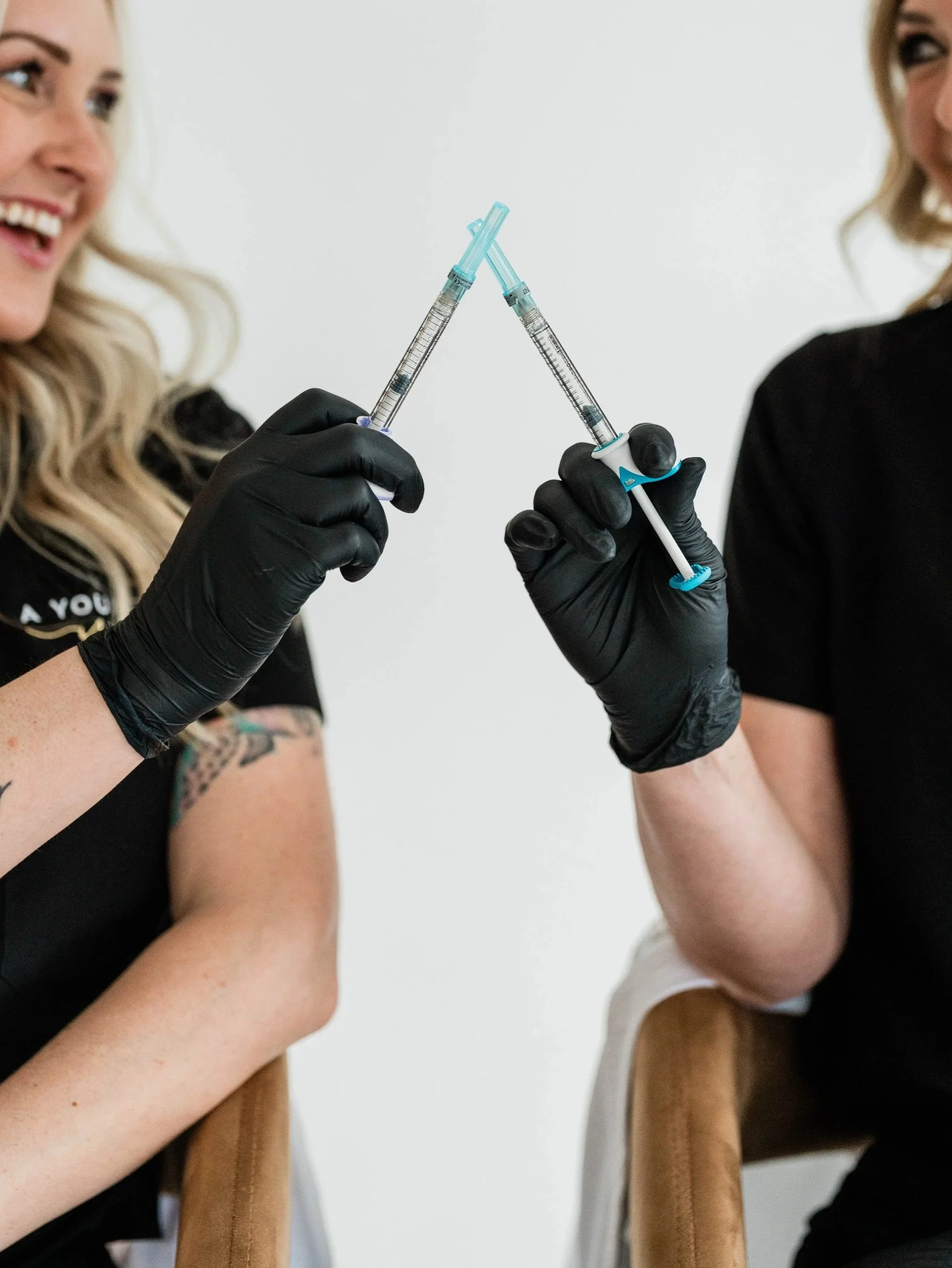 A Younger You Aesthetics nurse practitioners sitting next to each other toastiing syringes used for botox and other beauty fillers and injectables in a celevratory manner.