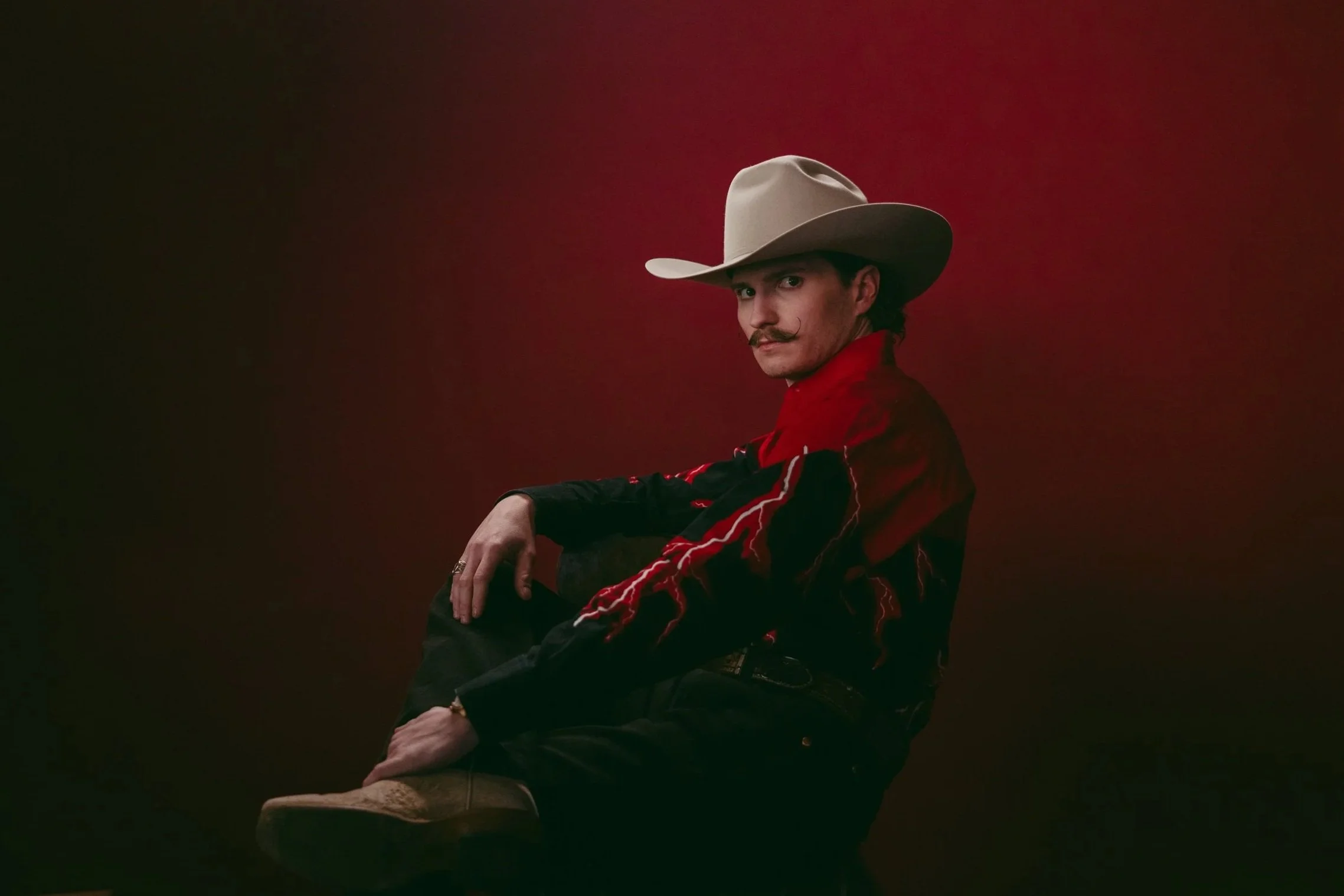 A man wearing a cowboy hat, a red and black jacket, and cowboy boots, sitting against a red gradient background, looking at the camera.