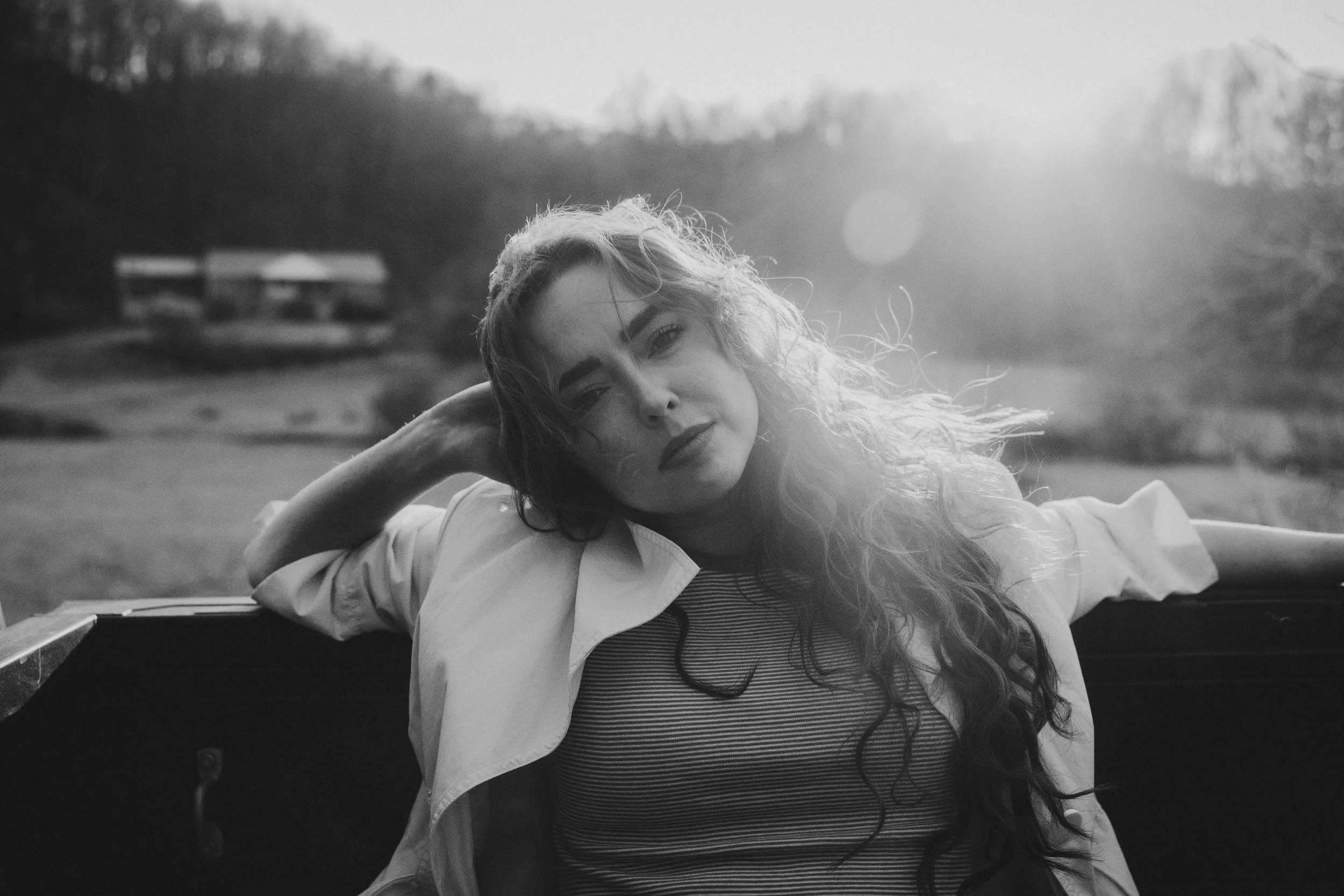 A young woman with long, wavy hair lying on her side outdoors, resting her head on her hand, with a serene expression on her face. The background shows a rural landscape with trees and a small building, illuminated by soft, natural light.