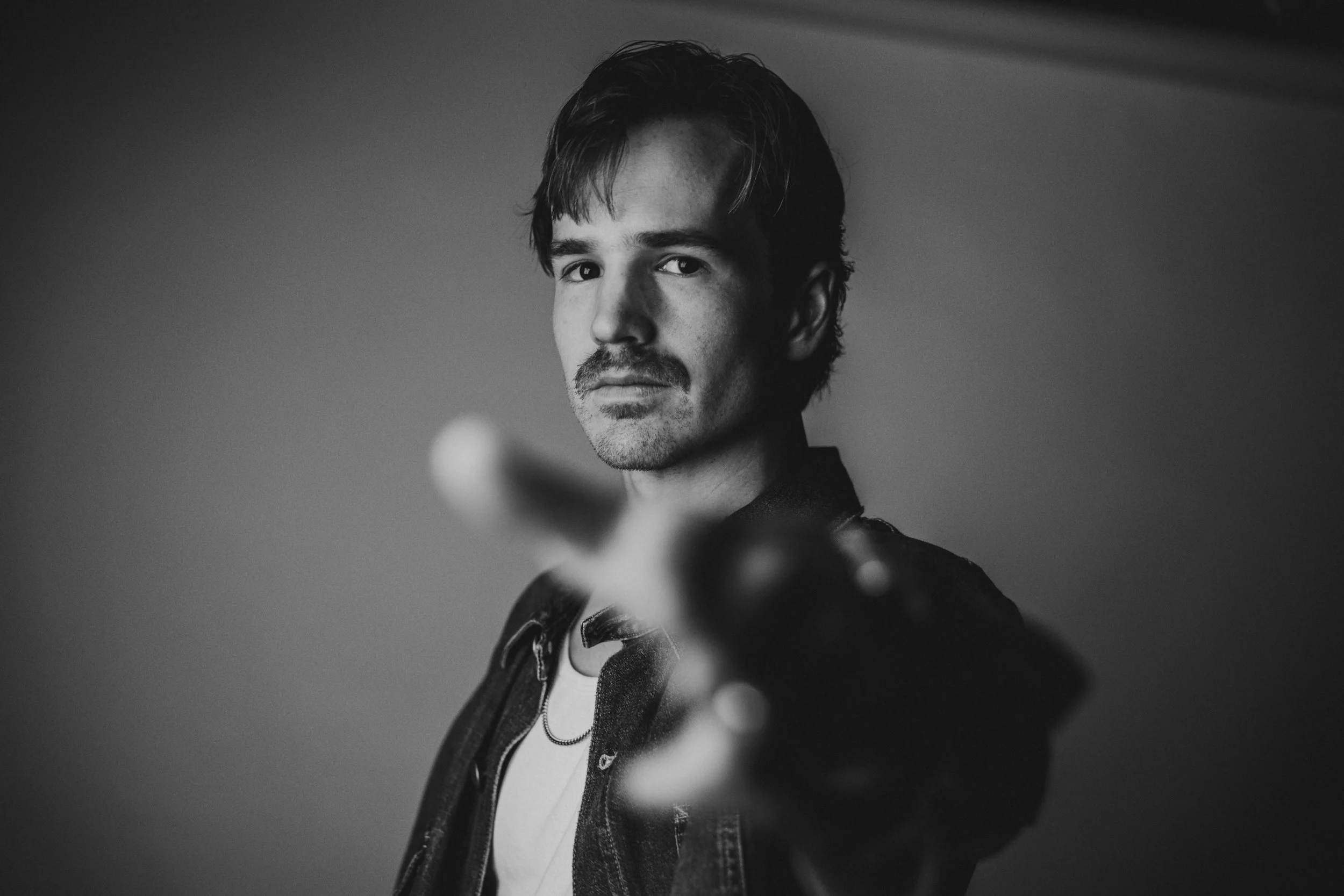 Black and white portrait of a young man with dark hair and a mustache, looking at the camera, standing against a plain background, with his hand extended towards the camera.