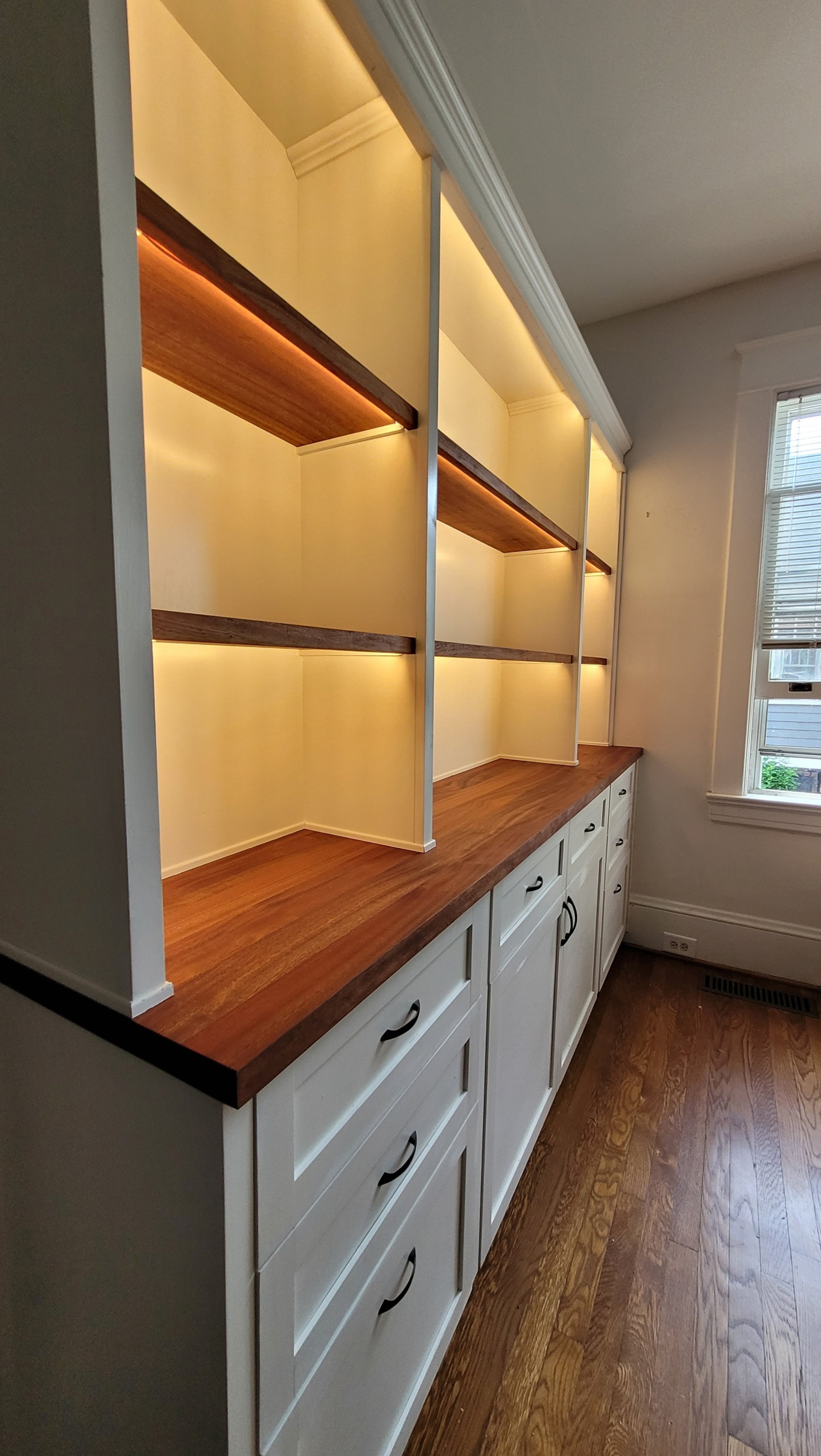 Built-in wooden cabinet with shelving and drawers in a room with hardwood floors and window blinds.