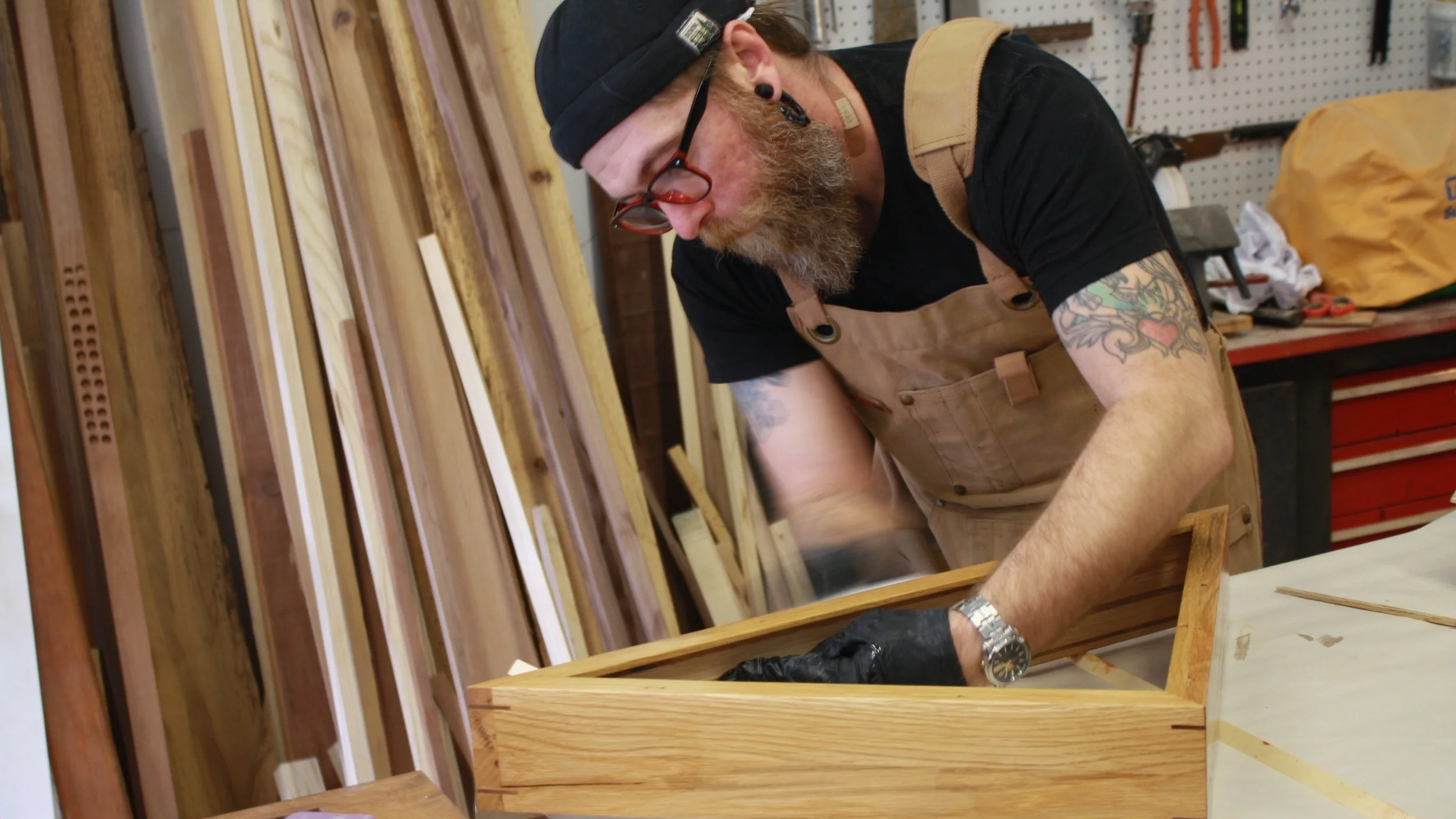 A man in a black shirt, black cap, and beige apron working on a wooden frame in a workshop filled with wood and tools.