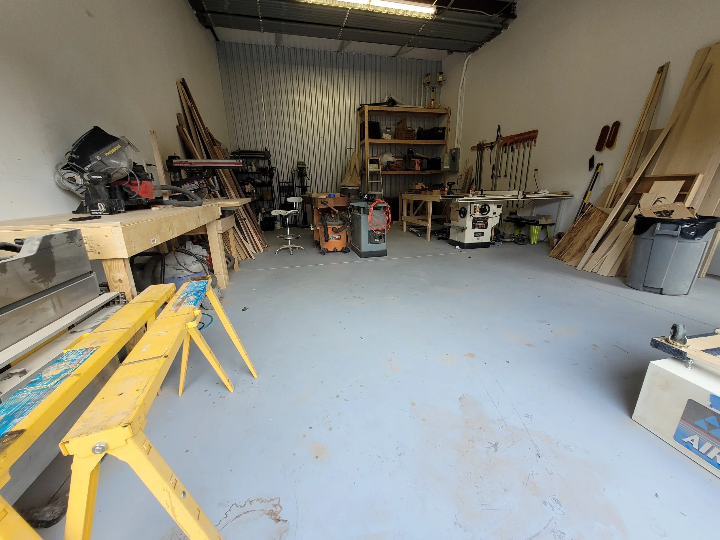 A spacious woodworking workshop with various tools, workbenches, and wood pieces; a yellow sawhorse in the foreground, shelves with equipment at the back, and a trash bin on the right.