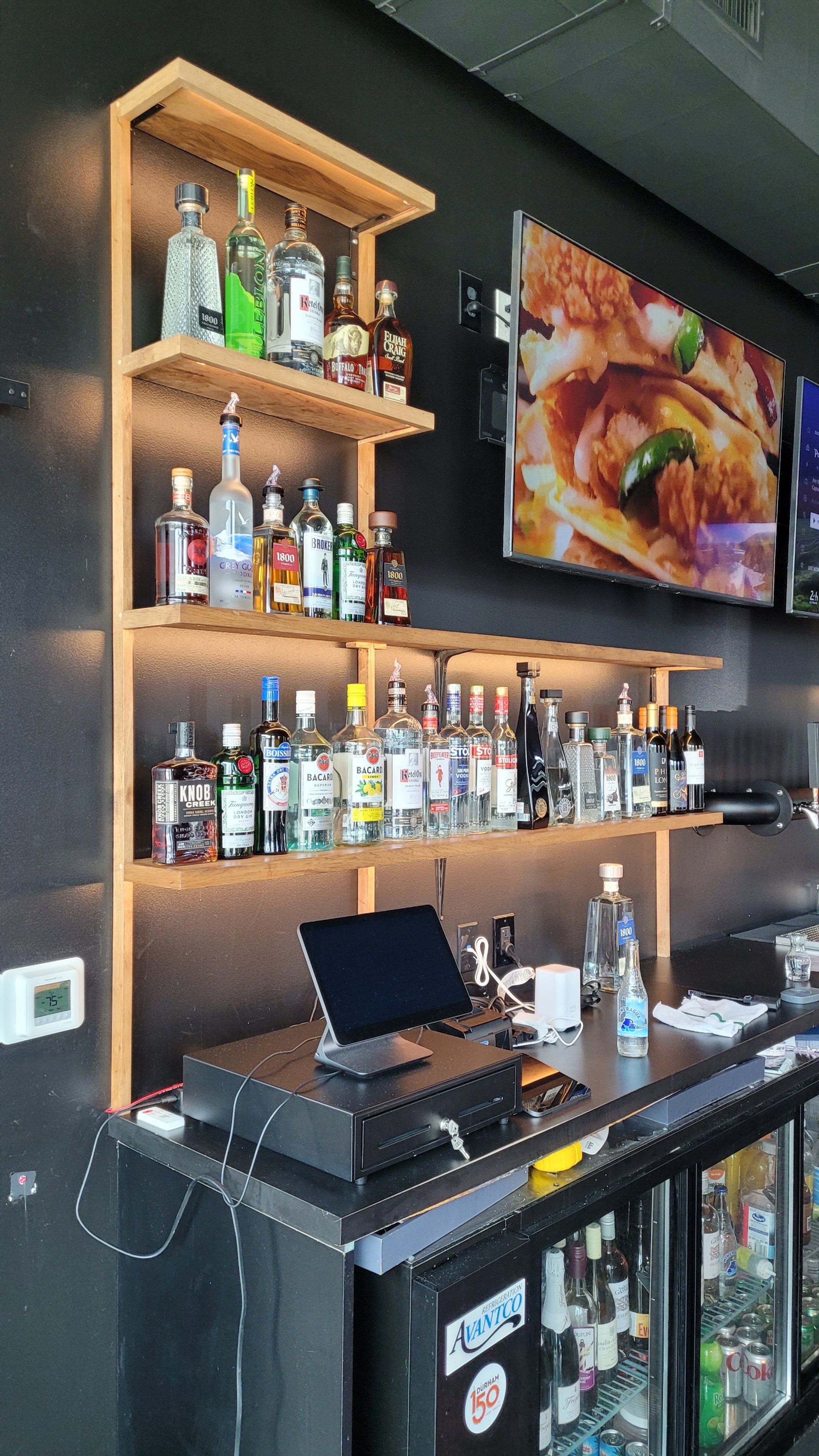 A bar setup featuring three wooden shelves stocked with various bottles of alcohol, a black countertop with a monitor, a cash register, and a mini fridge filled with beverages. A television screen displays a close-up of food, and a thermostat is mounted on the wall.